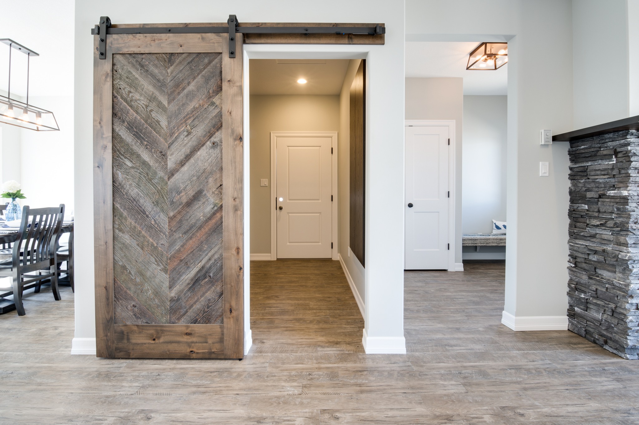 Modern interior with a rustic wooden sliding barn door revealing a hallway. Light wooden flooring, a dining area, and stone accent wall. Cozy ambiance.