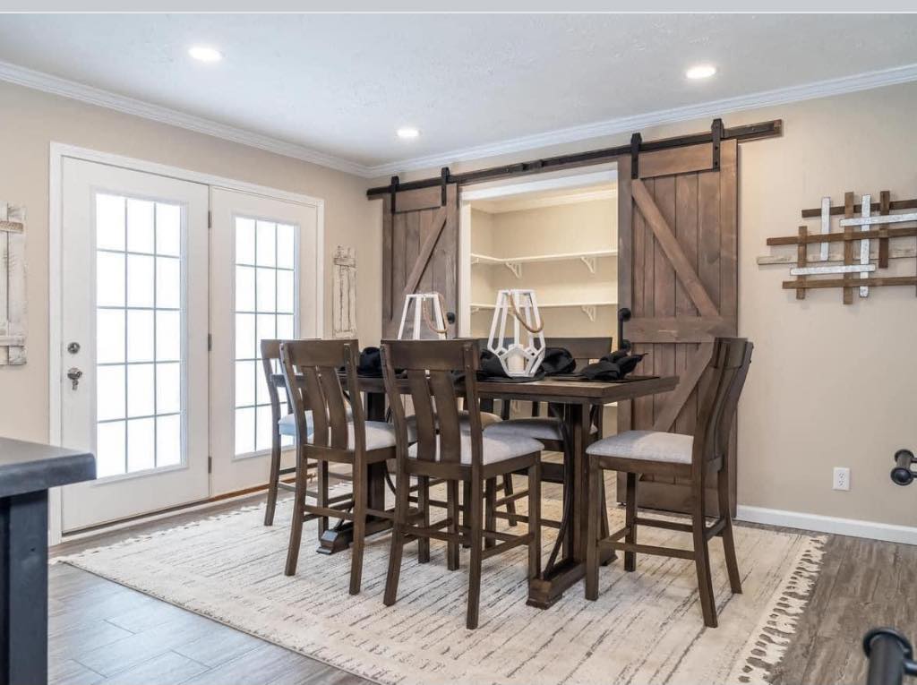 Dining room with a rustic wooden table and six chairs on a beige rug. Barn doors open to an alcove. French doors and abstract wall art add elegance.