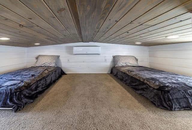 Loft with wooden ceiling and white walls, featuring low twin beds with dark gray bedding on each side. Soft carpet floor enhances cozy ambiance.