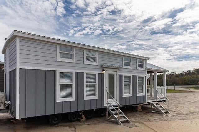 A modern gray and white tiny house on wheels under a partly cloudy sky. Two small staircases lead up to the door, conveying a cozy, portable home vibe.
