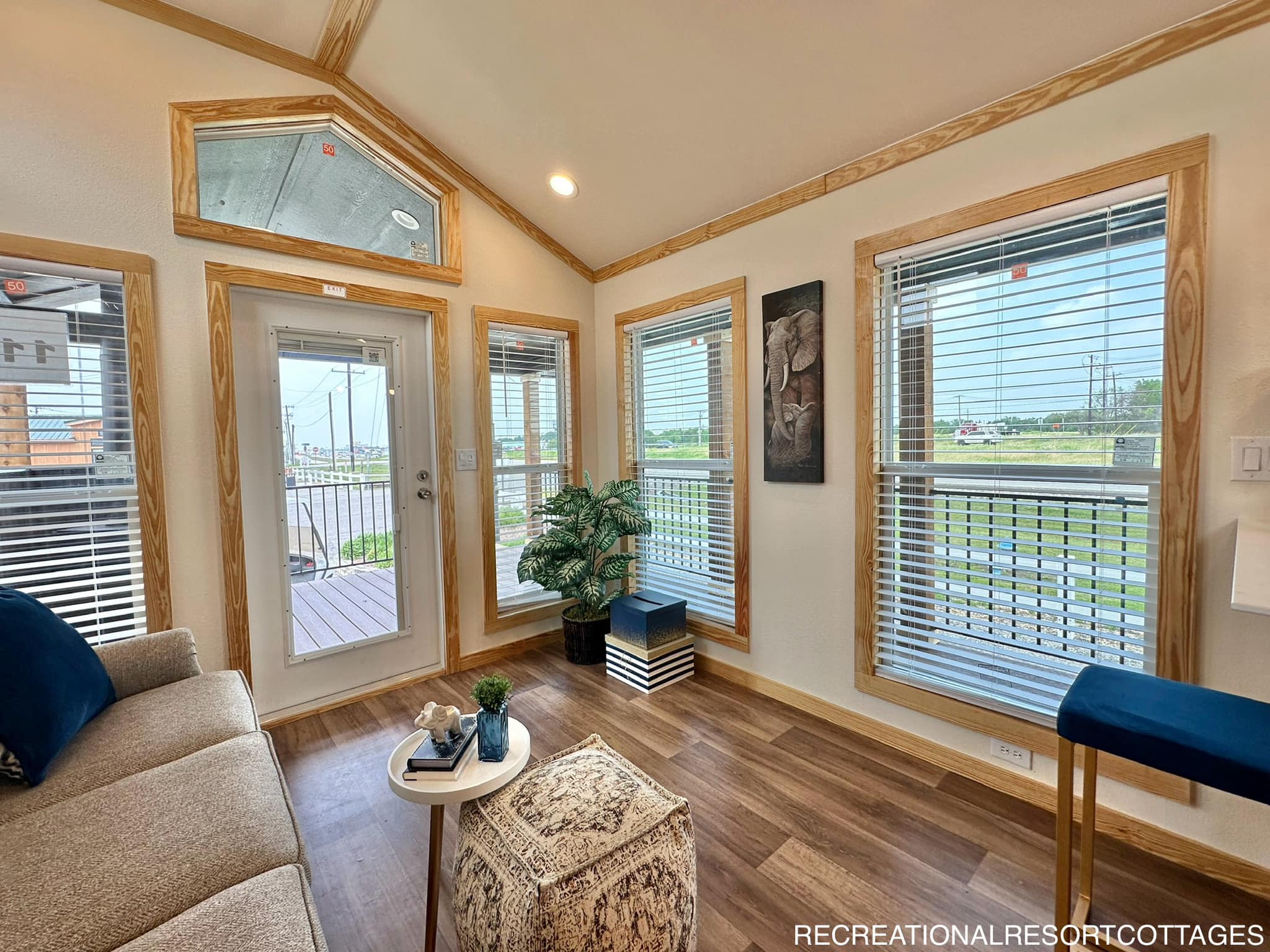 Cozy living space with large windows framed in wood, allowing natural light. Features a gray couch, blue accents, and potted plant, creating a calm vibe.