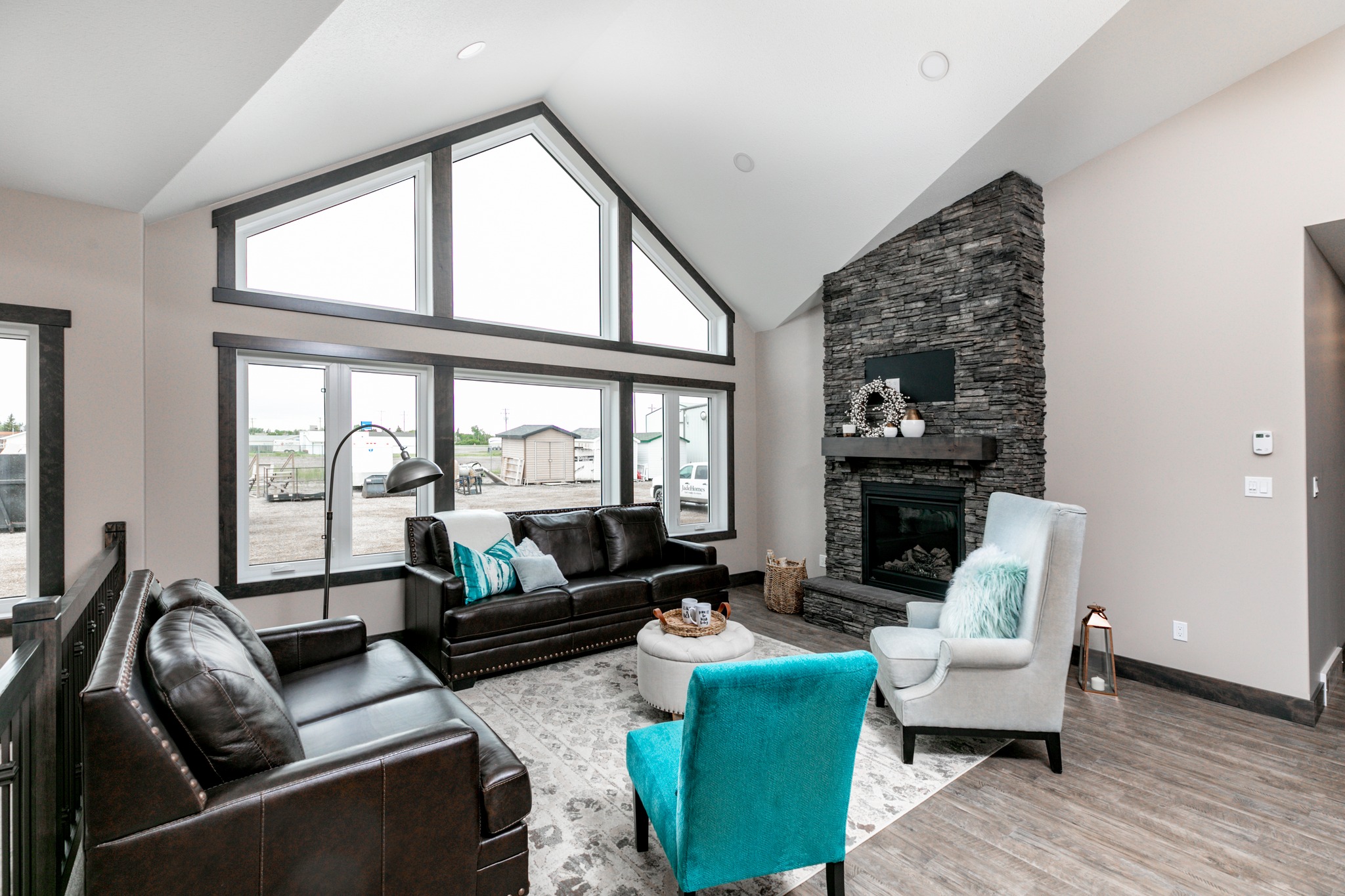 Modern living room with a high ceiling and large triangular windows. Features dark leather sofas, a stone fireplace, blue accents, and a round ottoman. Cozy and inviting.