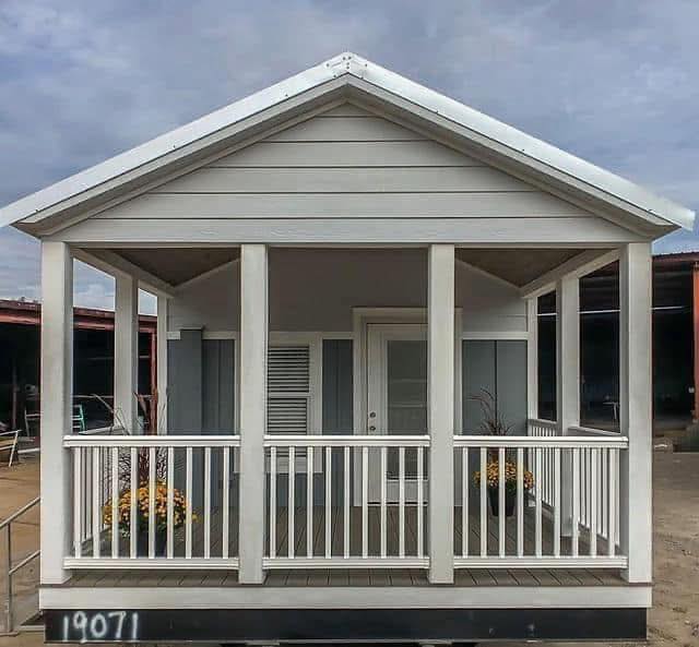 Small gray and white tiny house with a front porch featuring two potted plants and white railings, set against a cloudy sky, conveying a cozy feel.