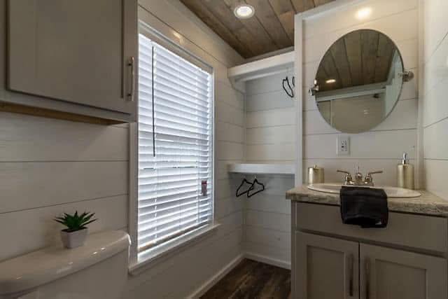 Cozy bathroom with wood accents, featuring a round mirror above a dual faucet sink, horizontal white shiplap walls, and natural light from window blinds.