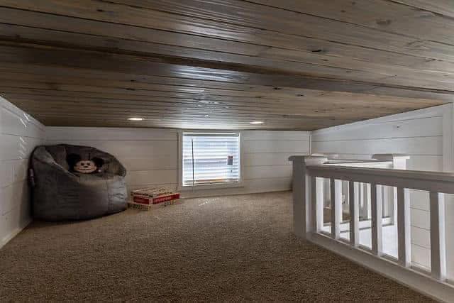 Cozy attic space with wood-paneled ceiling and beige carpet. There's a large bean bag with a stuffed animal, a window with blinds, and a toy on the floor near a railing.