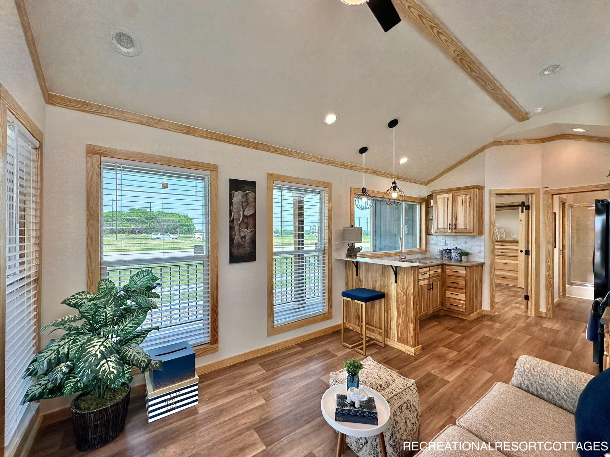 Elegant wooden interior of a cozy cottage living area with large windows, a kitchen with bar stools, indoor plant, and comfortable seating. Bright and inviting.