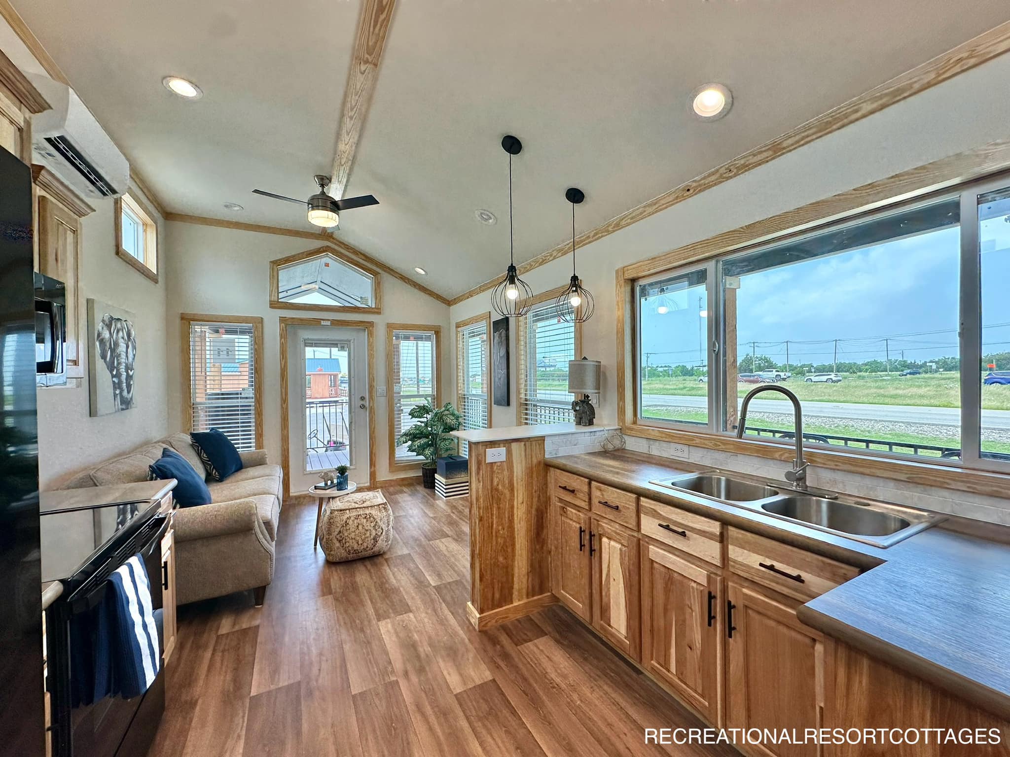 A cozy, modern tiny home interior with wooden floors and ceilings. A sofa and pouf sit near a large window. A kitchen area has pendant lights, cabinets, and a view outside, conveying warmth and comfort.
