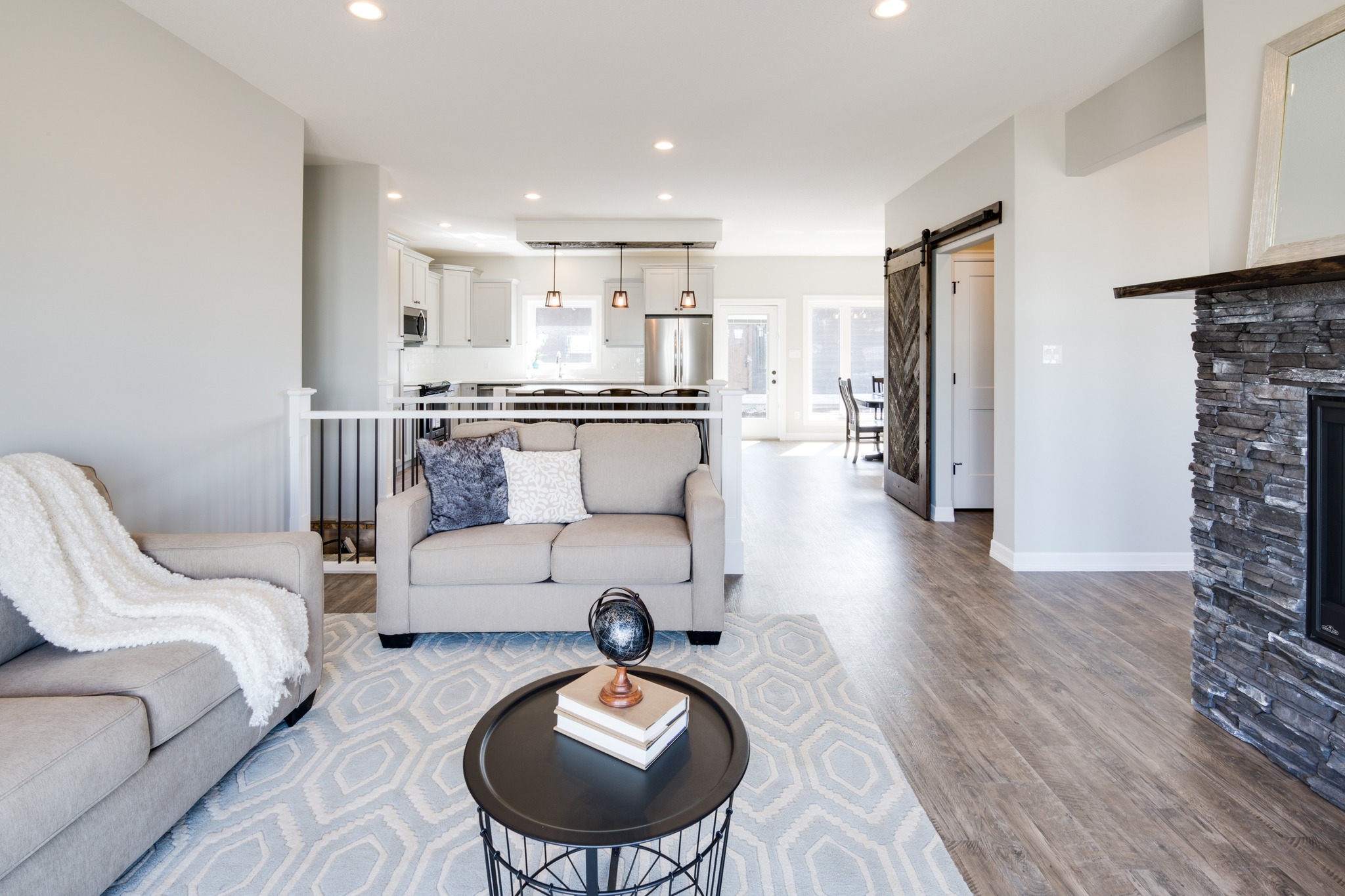 Spacious modern living room with neutral tones, featuring a beige sofa, geometric rug, and stone fireplace. Open kitchen visible, creating an inviting atmosphere.