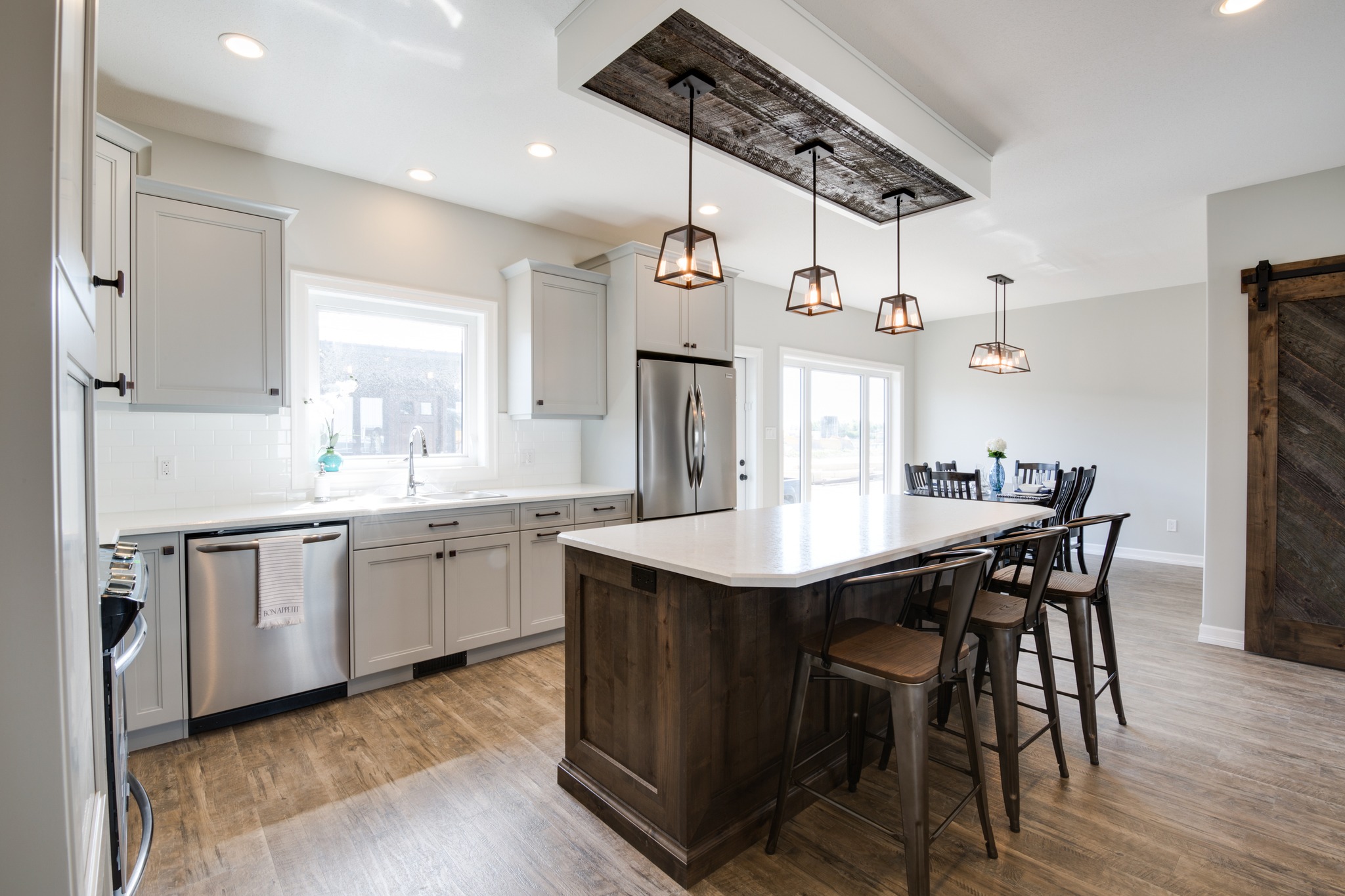 Modern kitchen with a central island, wooden bar stools, and pendant lights. Cabinets are white, and appliances are stainless steel, creating a warm, inviting tone.