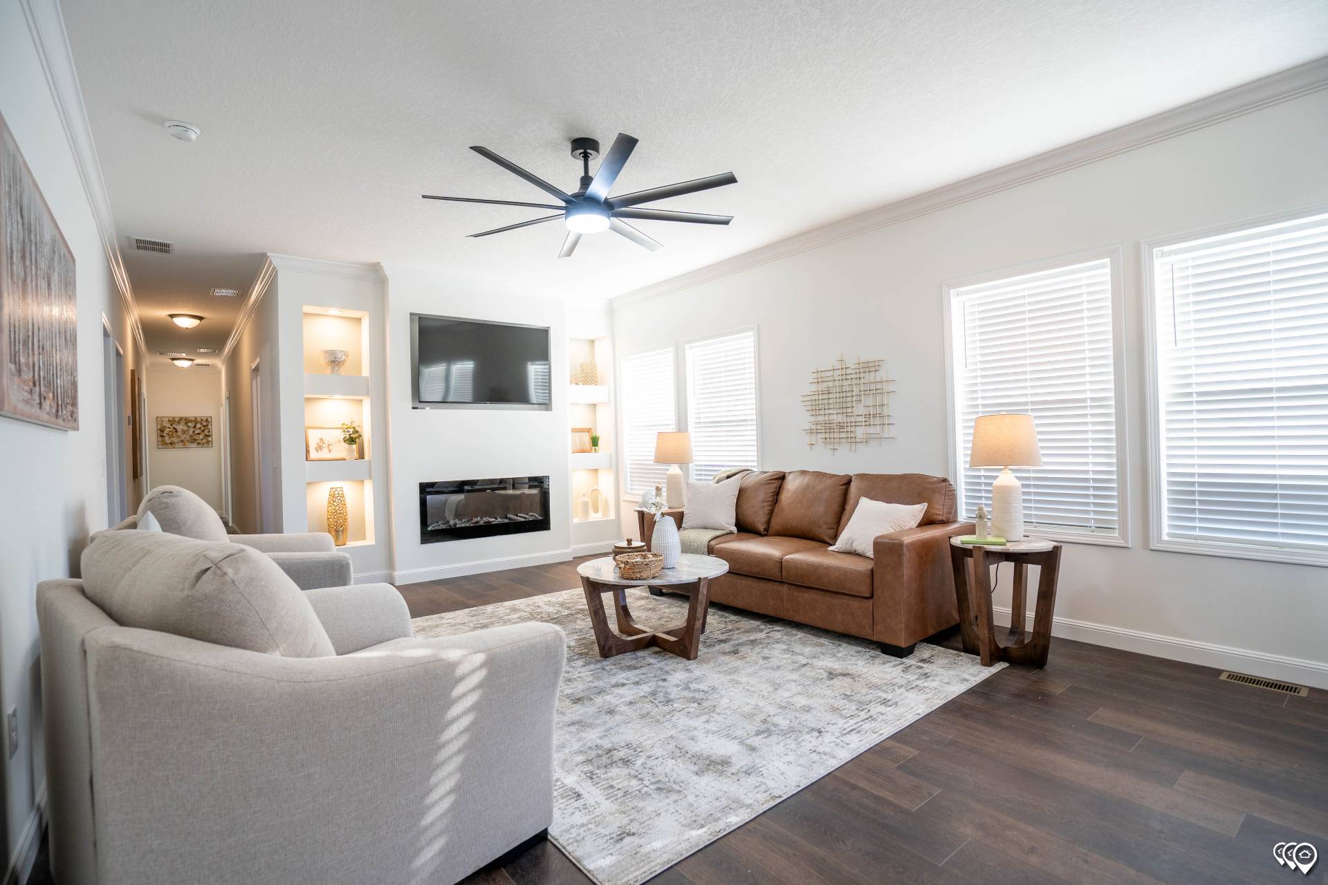 A modern living room with a brown sofa, white armchairs, and a coffee table on a gray rug. A fireplace and TV are on the wall, with shelves holding decor. Large windows bring in light.