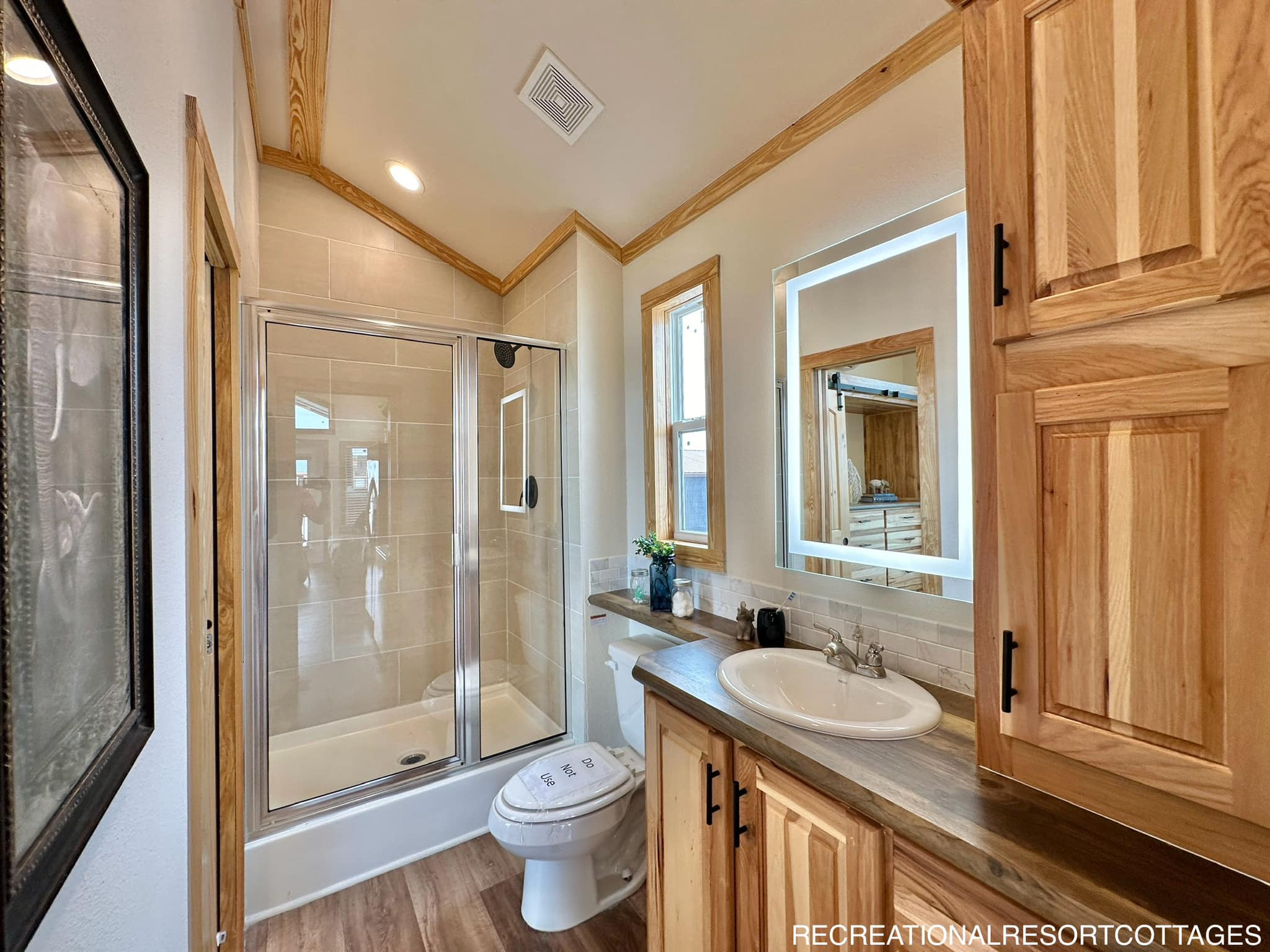 Compact bathroom featuring wood cabinetry, a glass-enclosed shower, illuminated mirror, and single window. Bright, rustic, and modern design.