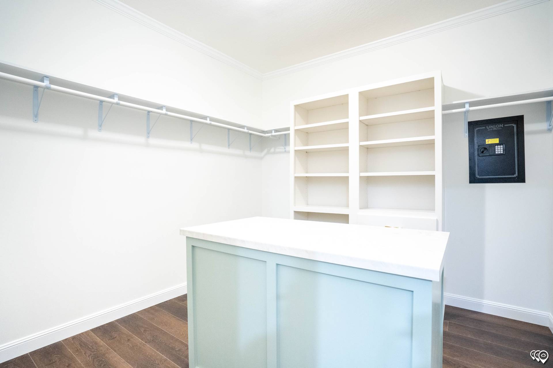 Bright walk-in closet with white walls and wood floor. Features open shelving, wall-mounted safe, hanging rods, and a central island with a white top.