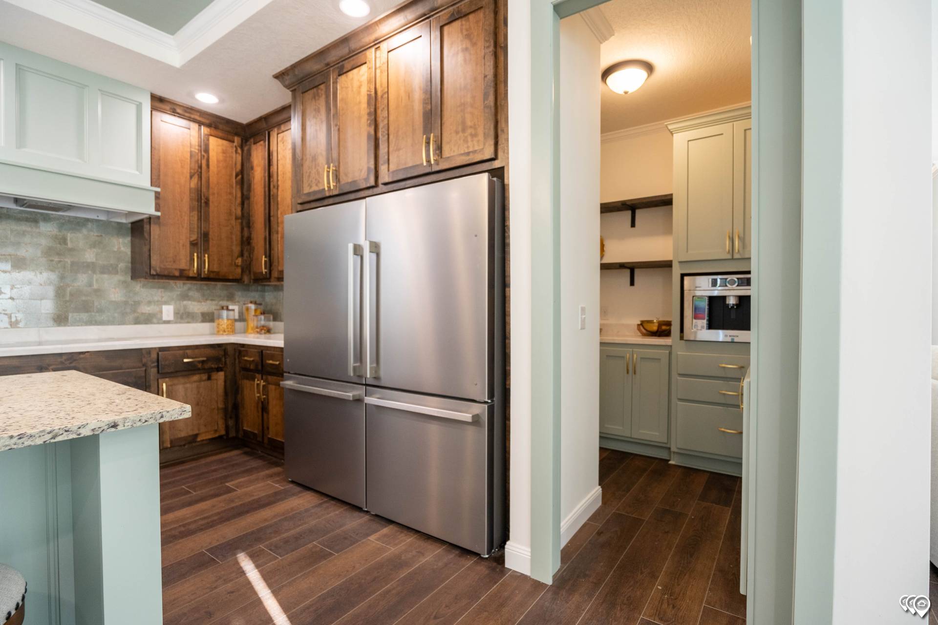 Airy kitchen with stainless steel fridge, wooden cabinets, granite counters, and wood flooring. Soft lighting creates a warm, inviting atmosphere.