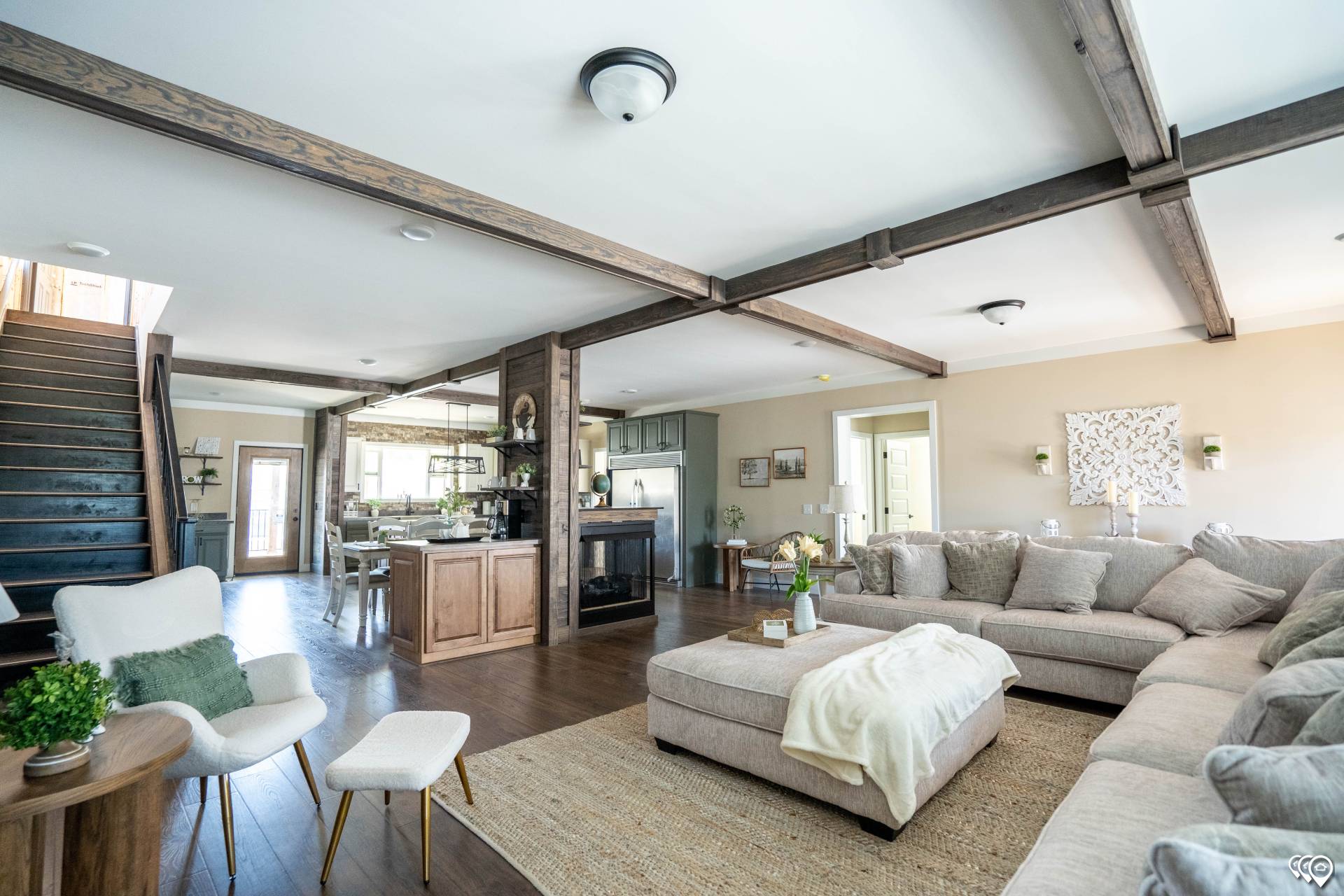 Spacious living room with neutral tones, featuring a large sectional sofa, ottoman, and cozy decor. Exposed wooden beams and an open kitchen are visible.