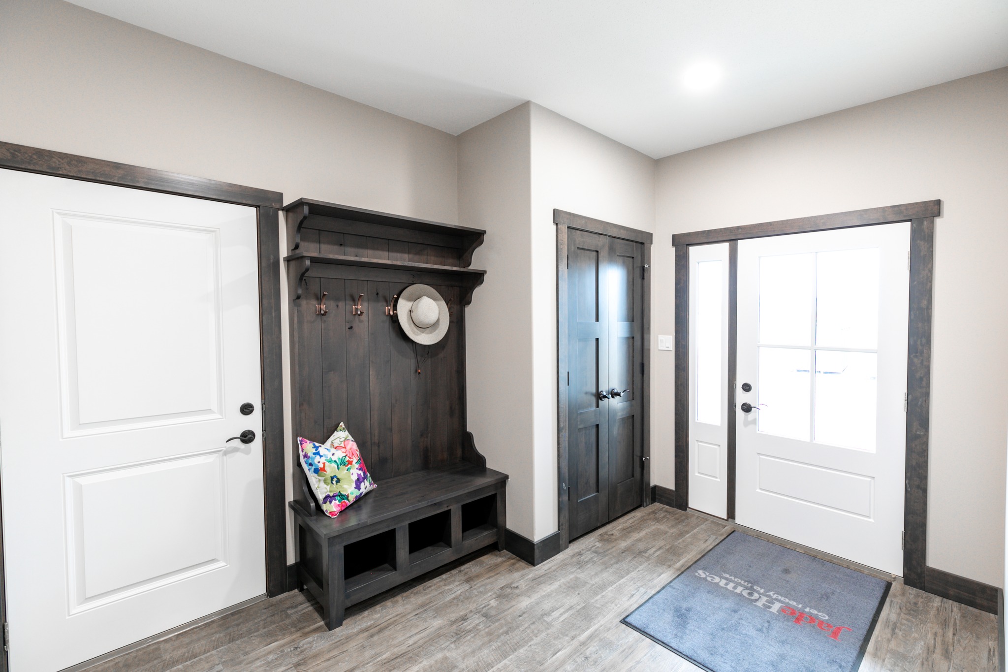 Bright entryway with light wood floor, dark wood bench with colorful pillow and hat on hooks. White doors have dark trim. Calm, welcoming atmosphere.