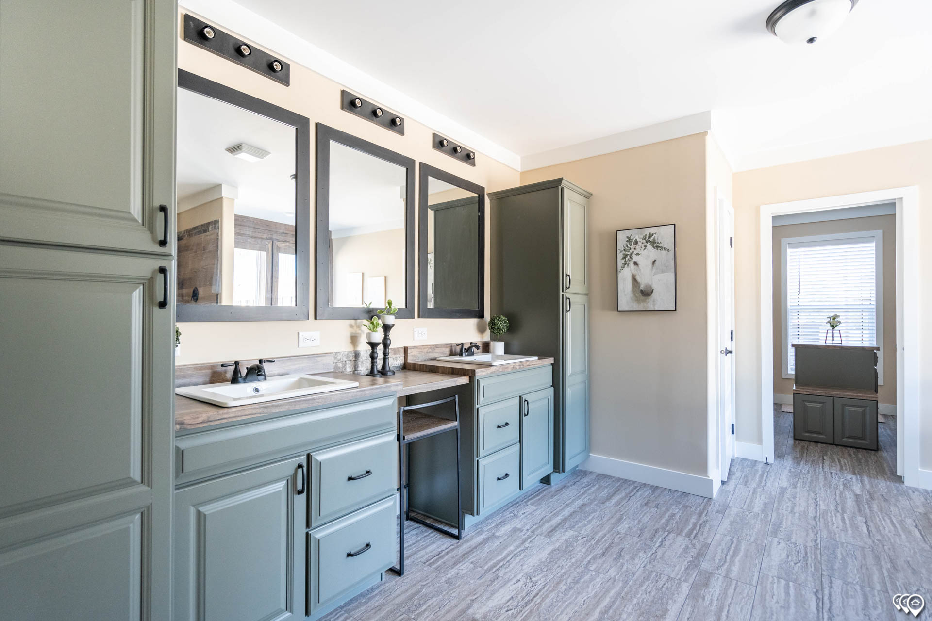 Spacious bathroom with sage green cabinets, dual sinks with mirrors above, taupe wall paint, and gray wood-textured tile flooring. A serene, bright atmosphere.