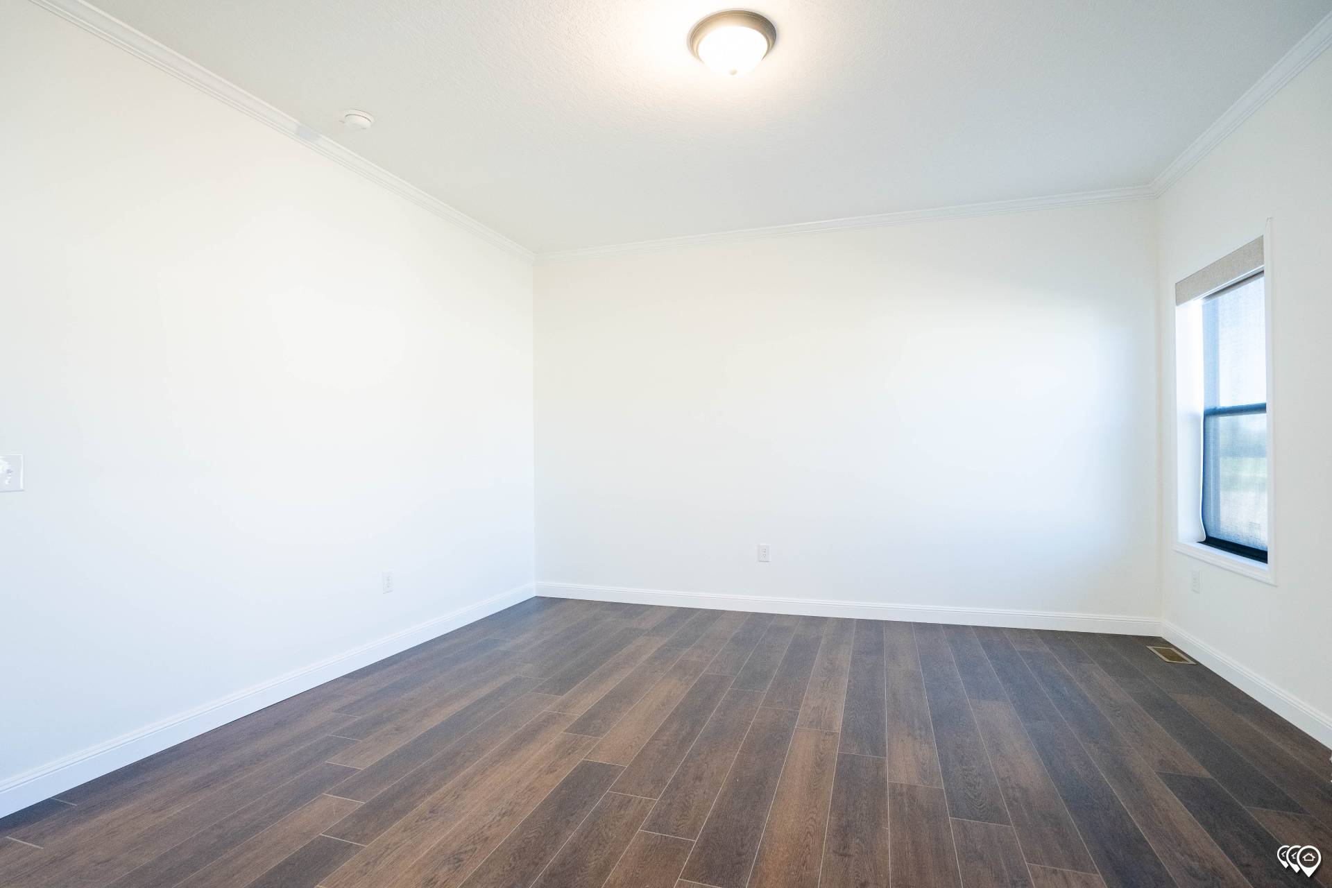 An empty room with white walls, a ceiling light, dark wood flooring, and a window on the right, creating a bright and clean atmosphere.