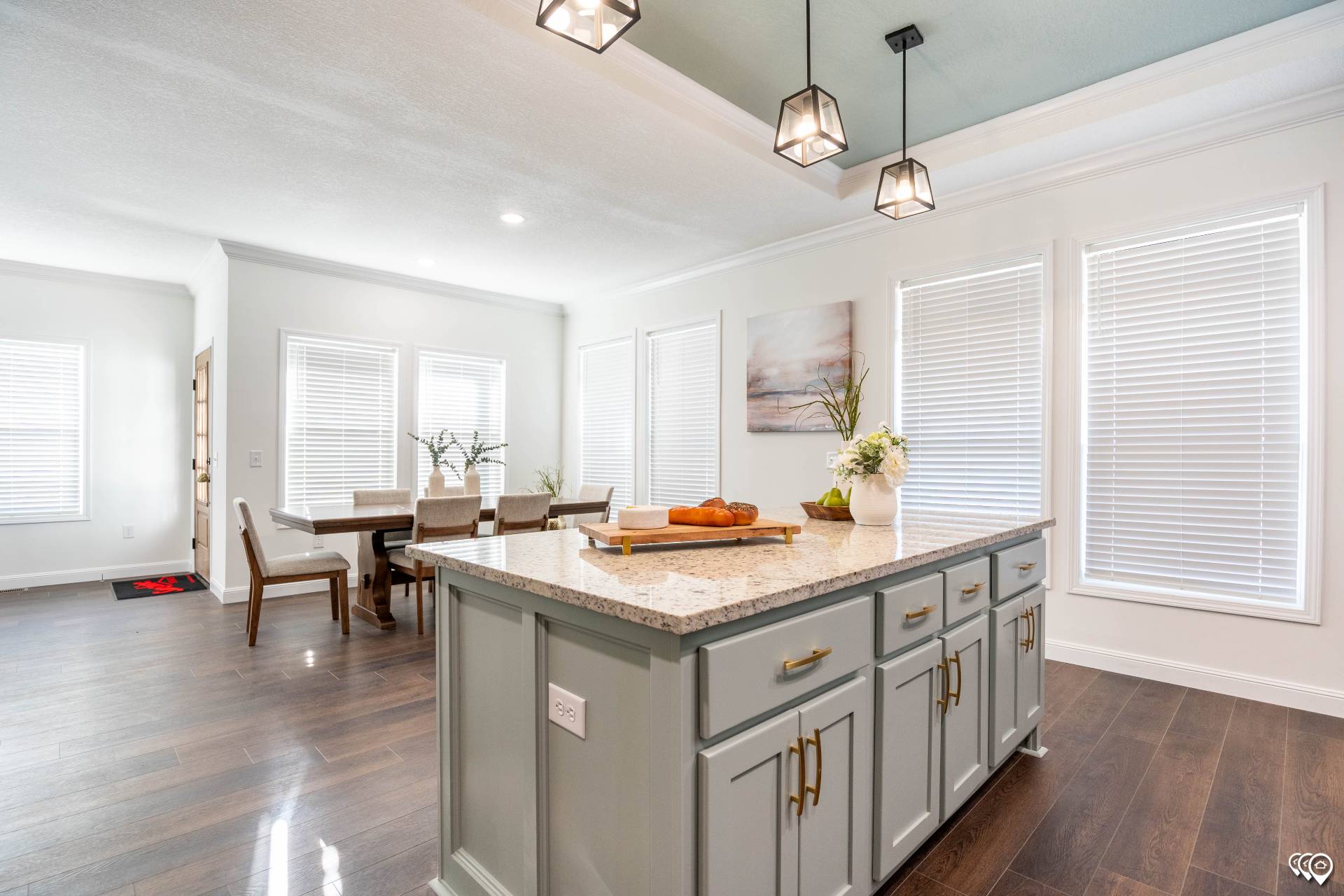 Bright kitchen interior with a granite island and teal cabinets. Pendant lights hang above. A dining table with chairs is in the back. Modern and inviting.