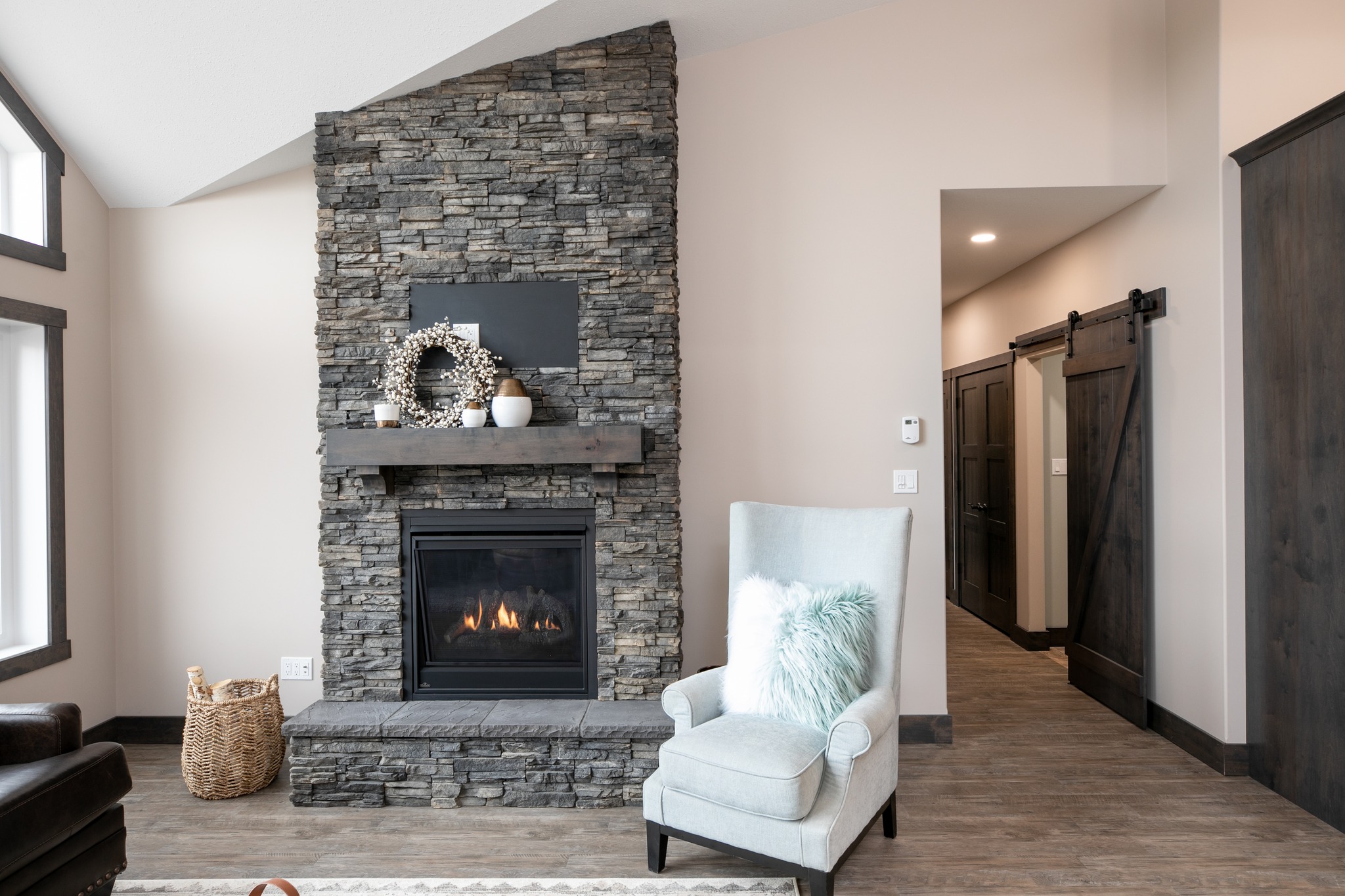 A cozy living room featuring a stone fireplace with a warm fire, a wooden mantle with decor, a plush armchair with a fur pillow, and nearby barn doors.