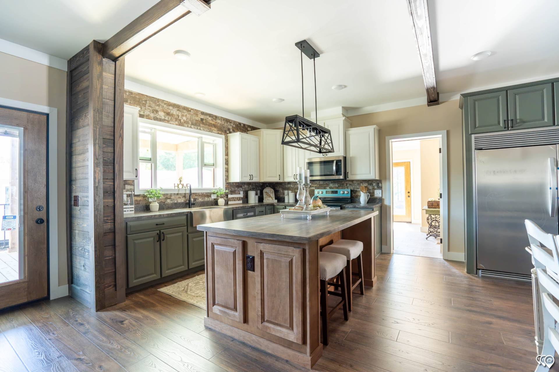 Spacious kitchen with rustic wooden beams, a central island with bar stools, stainless steel appliances, and sunlight streaming through large windows.