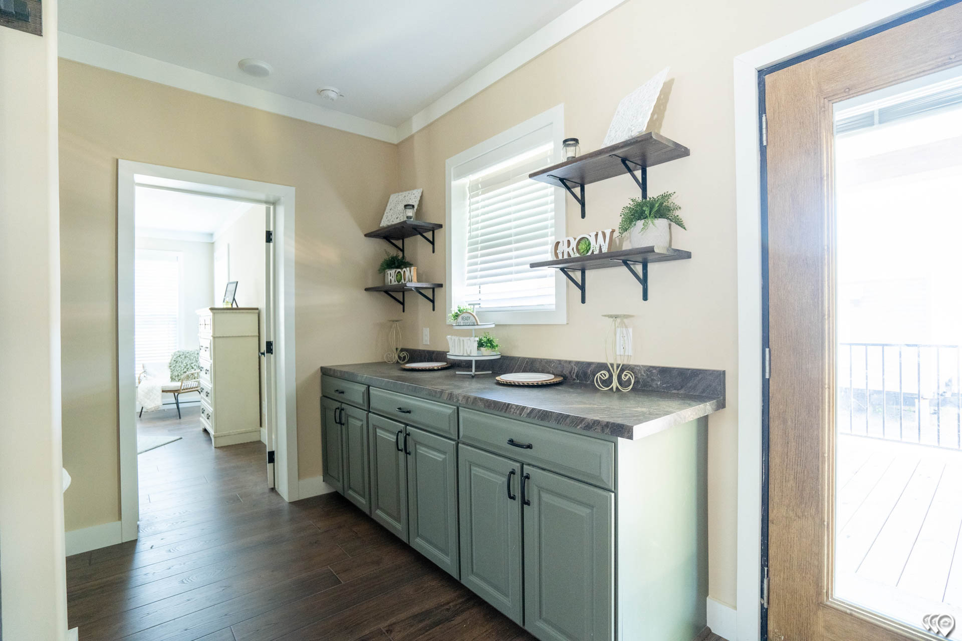 A bright, inviting hallway with light beige walls and dark wood floors. Green cabinets with a stone countertop are below three wall shelves, adorned with small decor and plants. A large window on the right lets in natural light, creating a warm ambiance. A doorway leads to a room with white furniture.