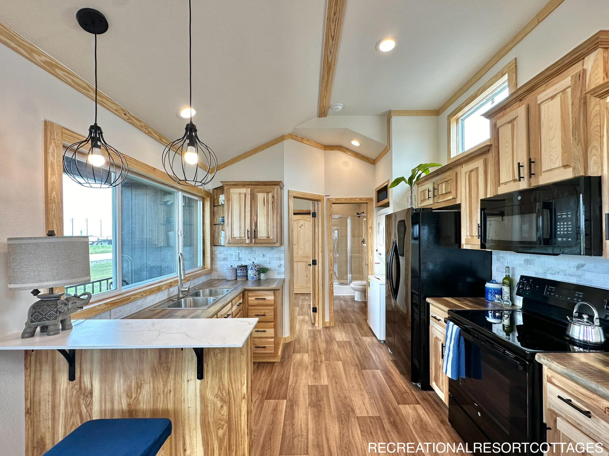 Modern kitchen with wooden cabinets and flooring, black appliances, and pendant lighting. A window offers natural light, creating an inviting atmosphere.