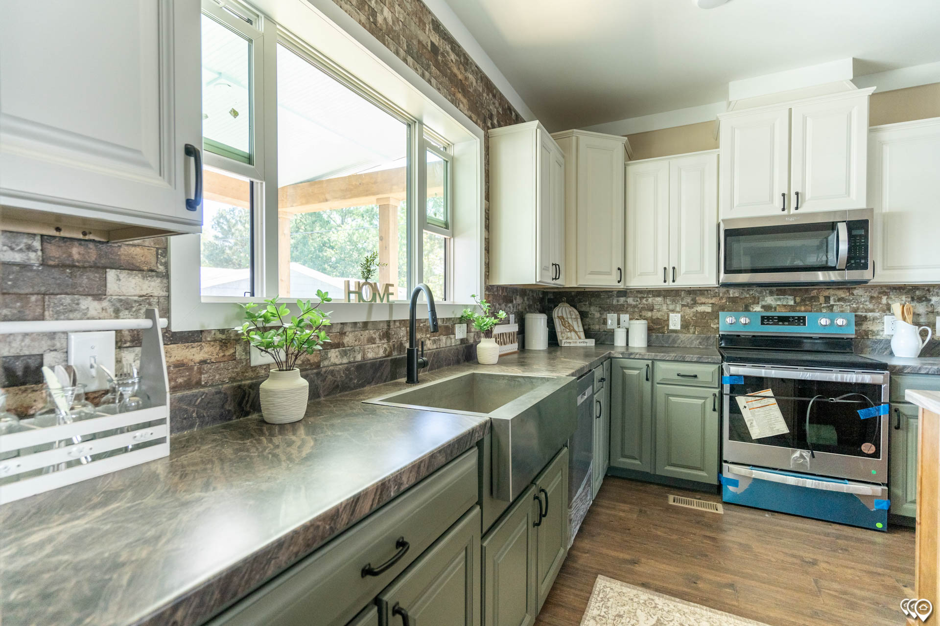 Bright kitchen with stainless steel appliances, white cabinets, and dark countertops. Large window, rustic brick backsplash, and plants add warmth.