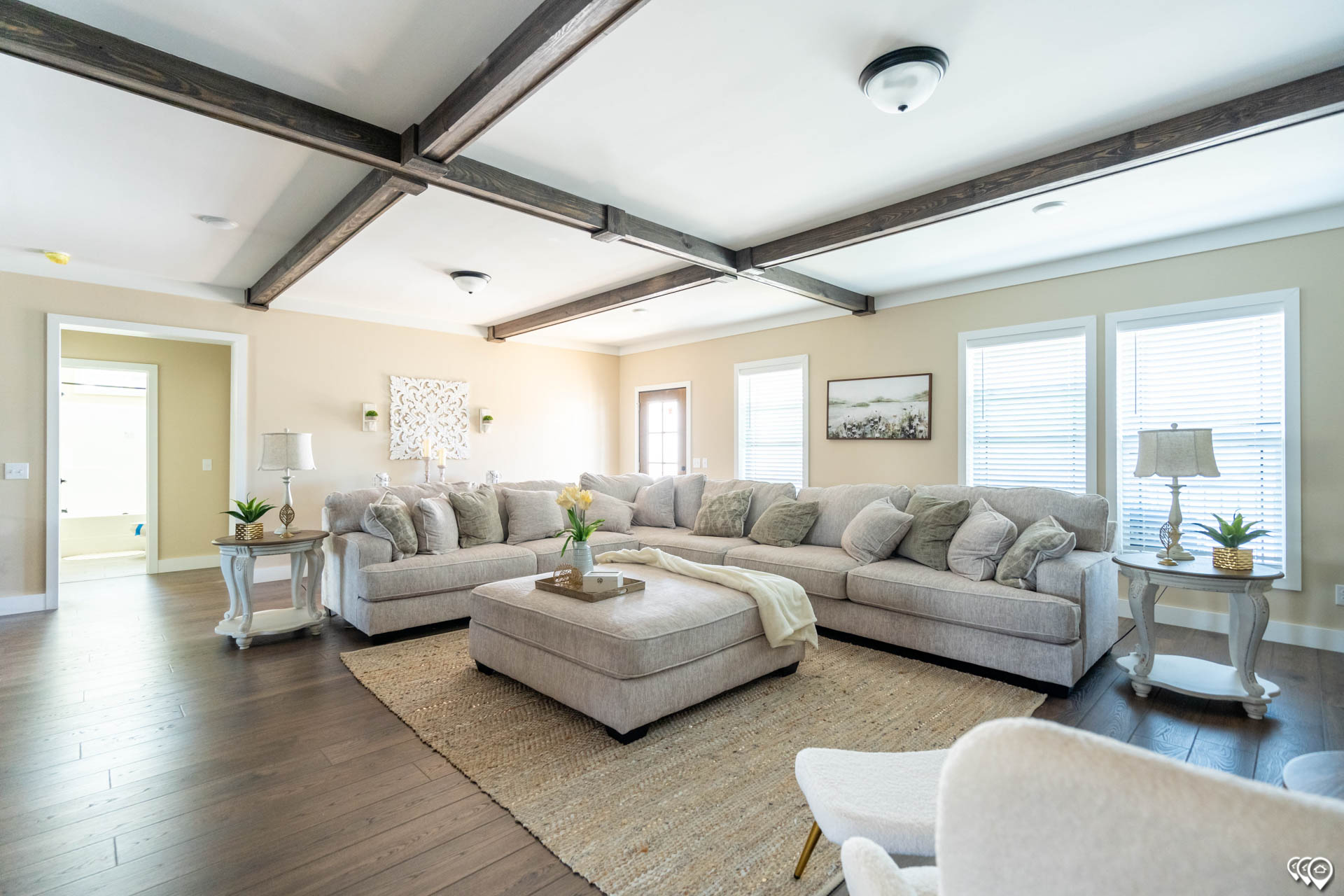 Spacious living room with a large L-shaped beige sectional sofa, light wood floors, and rustic beams. Soft lighting creates a warm, inviting atmosphere.