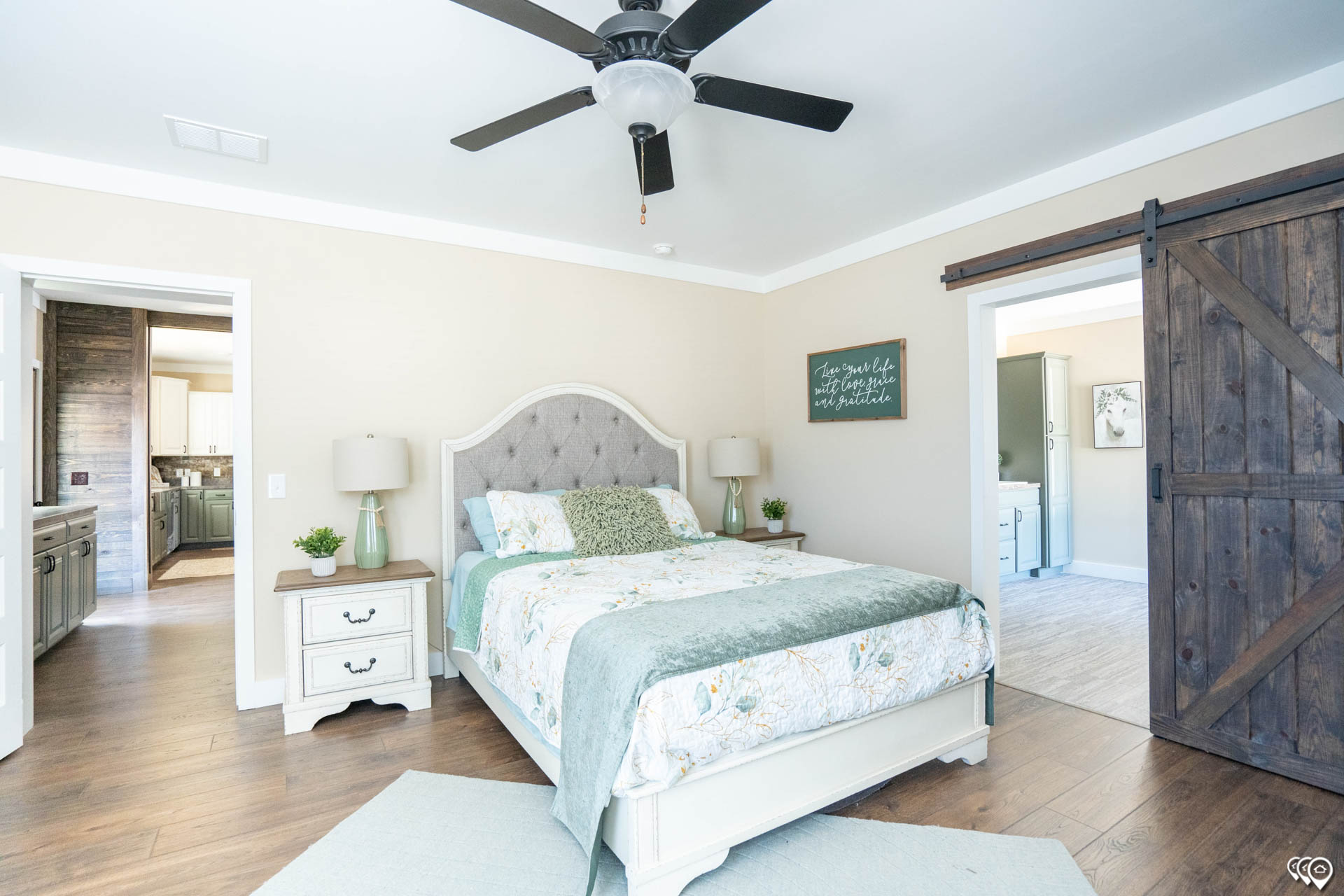 Spacious bedroom with a neutral palette, light floral bedding, and a tufted headboard. Barn door on right, open to a hall. Ceiling fan above. Cozy and airy.