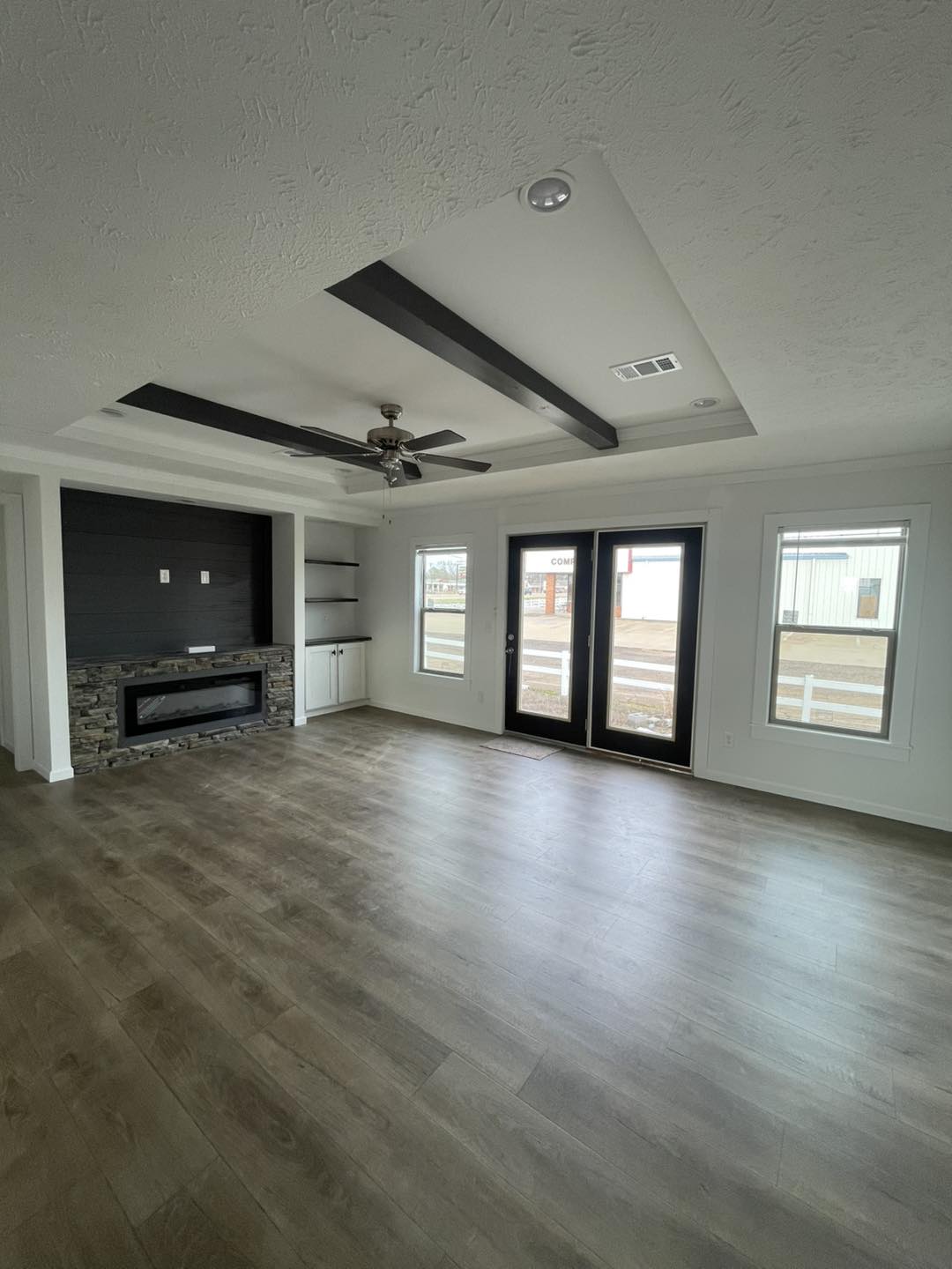 Spacious living room with light wood flooring, a modern stone fireplace, built-in shelves, and two large windows with French doors. Ceiling fan adds elegance.