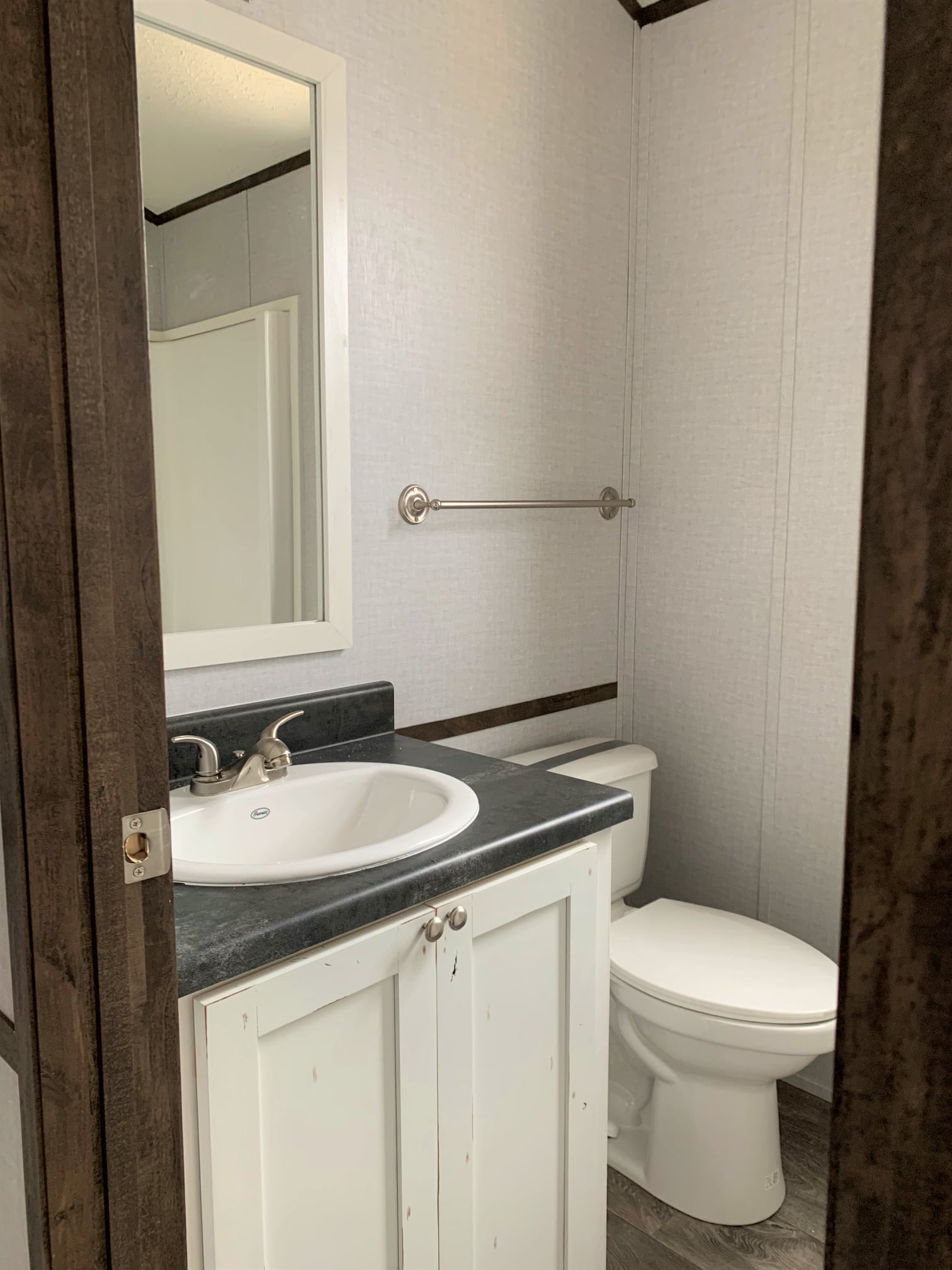 Small bathroom with a white sink on a dark countertop, a mirror, towel rack, and toilet. Light walls and rustic wood accents create a cozy feel.