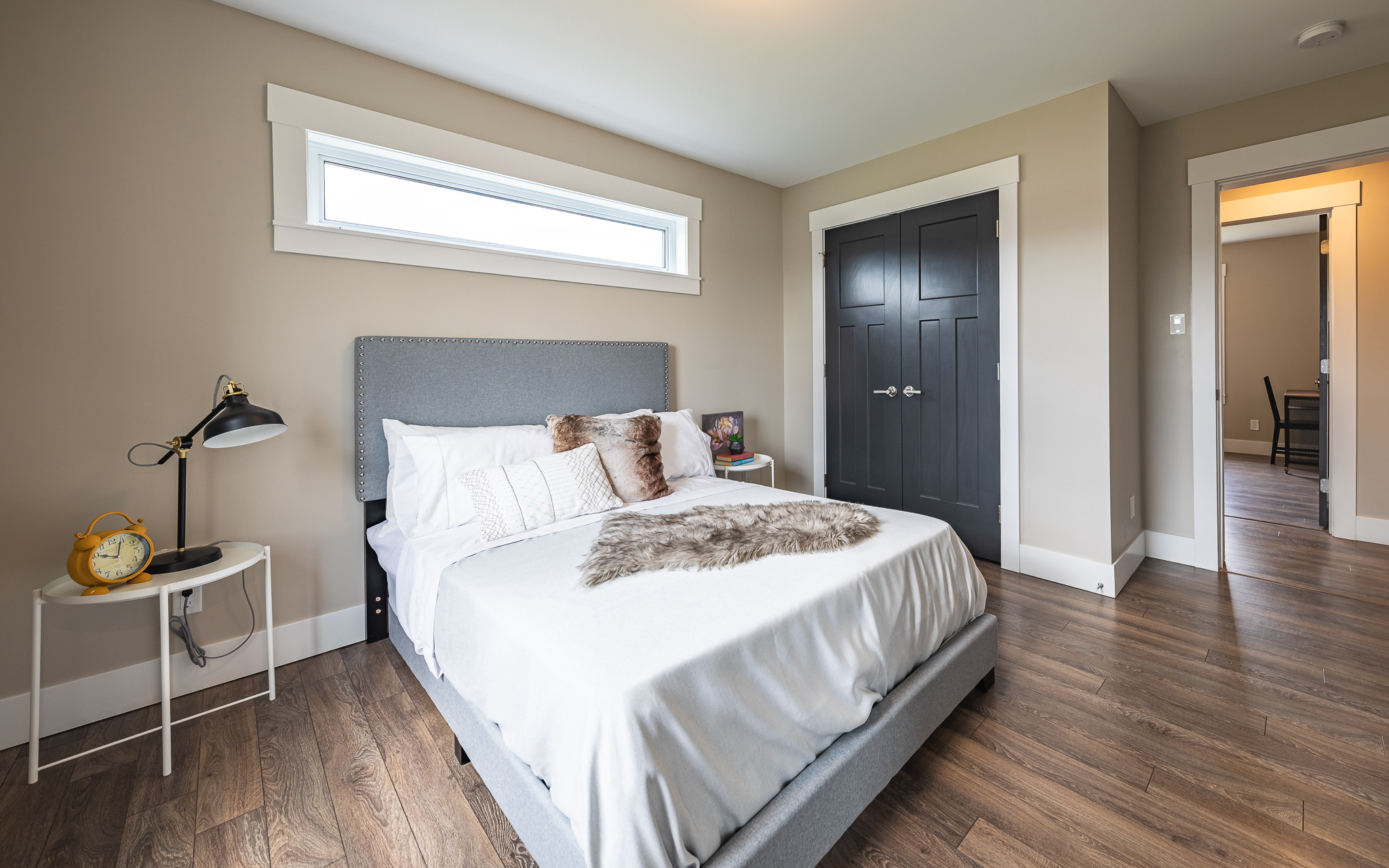 A cozy bedroom features a neatly made bed with white and brown pillows and a gray headboard. A fur throw adds texture. A black lamp and a yellow clock are on a white side table, complementing a modern, serene ambiance.
