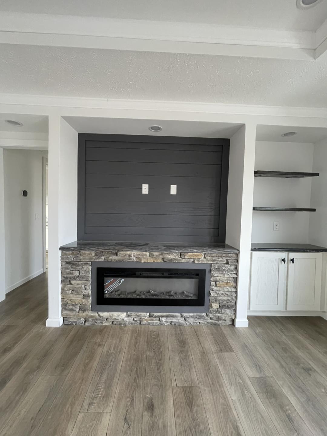 Living room with stone fireplace, wooden shelves, and white cabinetry. Gray accent wall adds contrast. A cozy, modern atmosphere is conveyed.