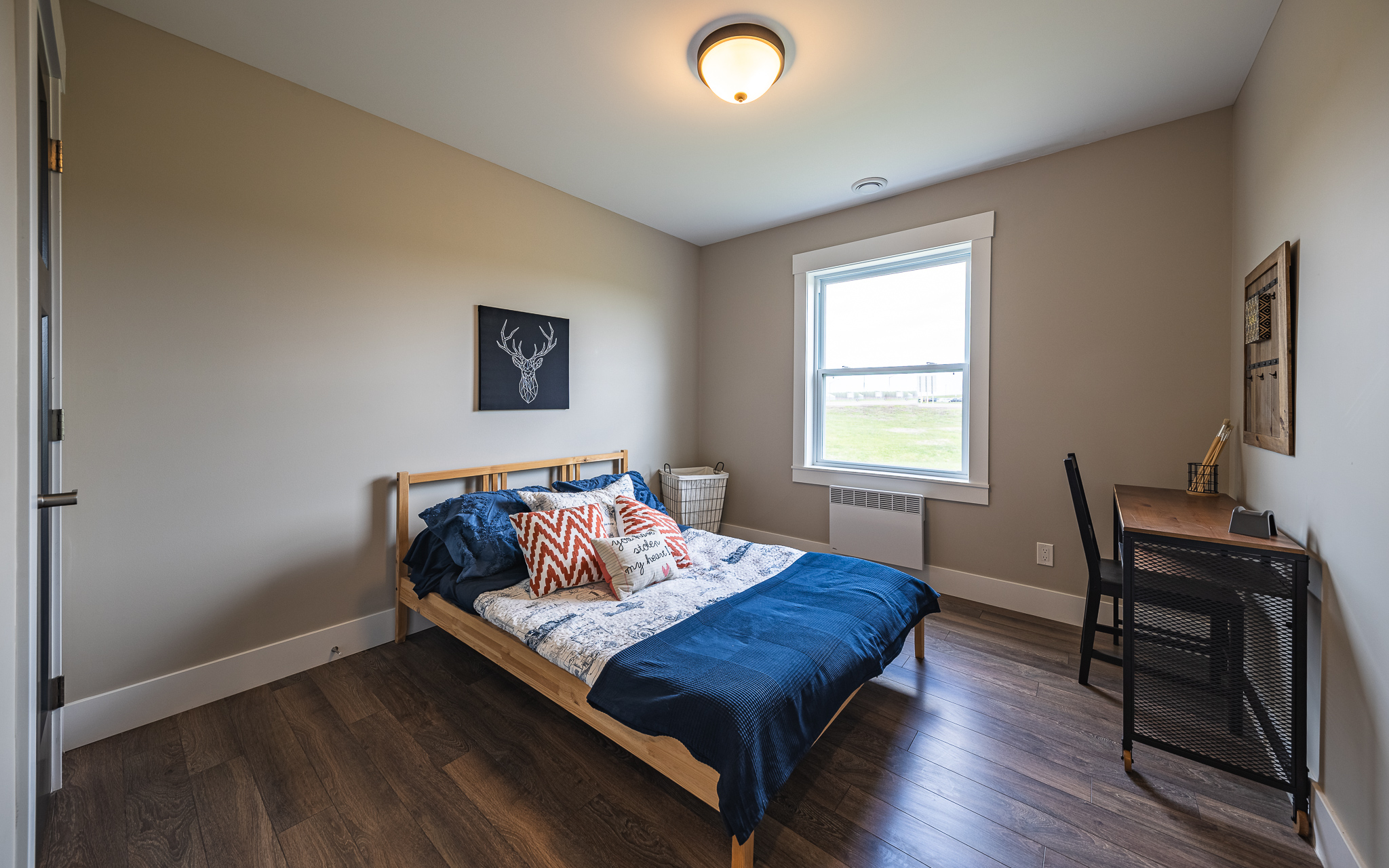A cozy bedroom with a wooden bed, adorned with a blue comforter and patterned pillows, a window showing a clear view outside, and a small desk and chair.