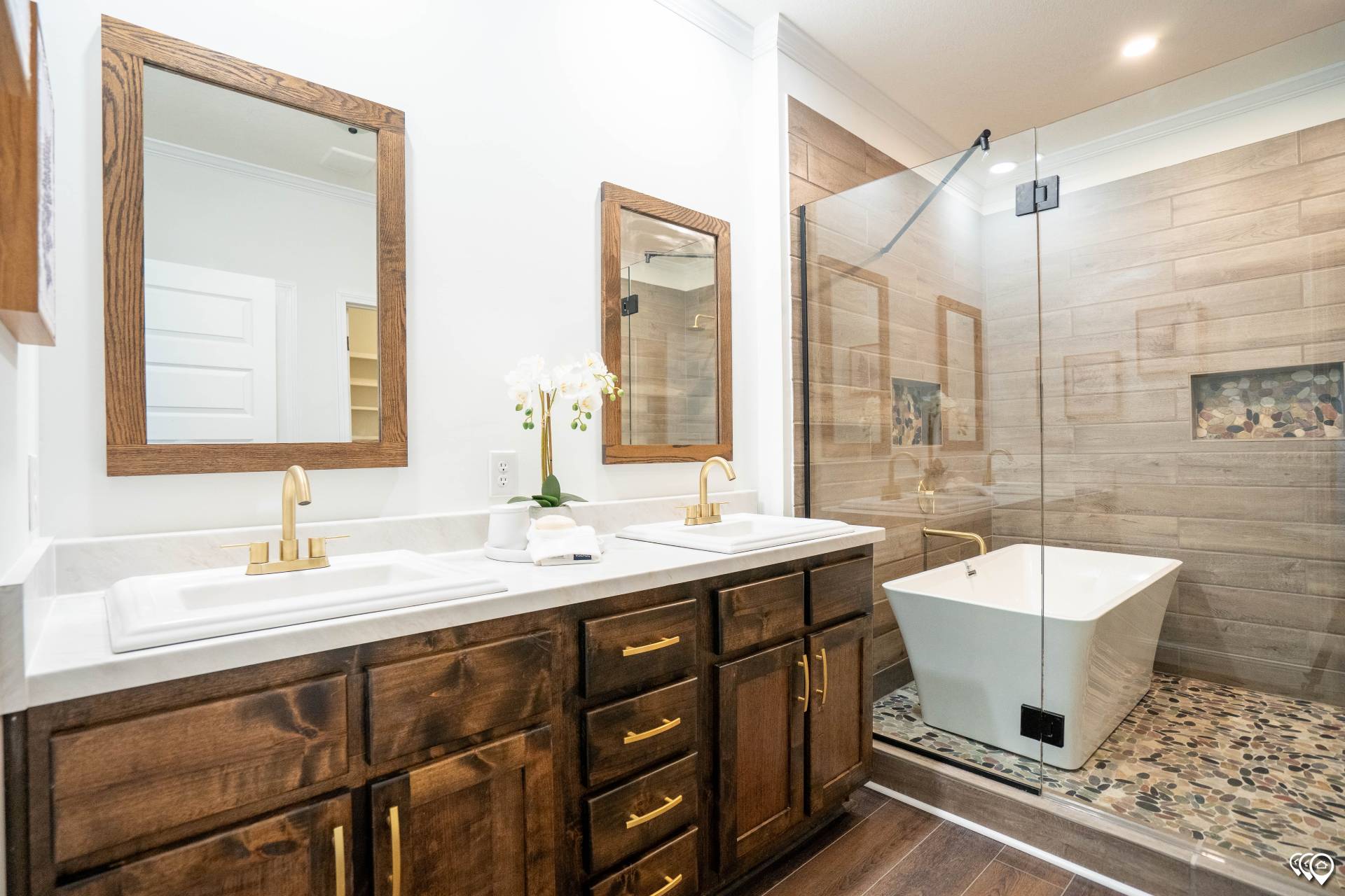 Modern bathroom with dual sinks on wooden vanity, gold faucets, large mirrors, glass-enclosed shower with stone floor, and a freestanding tub. Luxurious and serene.
