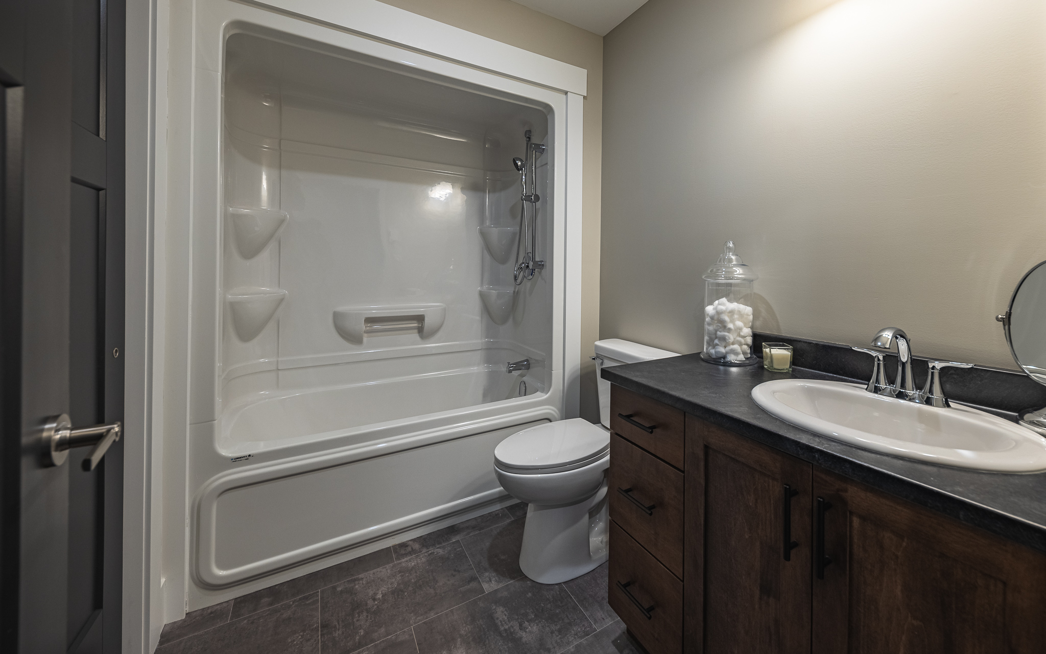 Modern bathroom with white bathtub-shower combo, gray tiles, wooden vanity with sink, and glass jar of cotton balls, creating a clean, inviting feel.