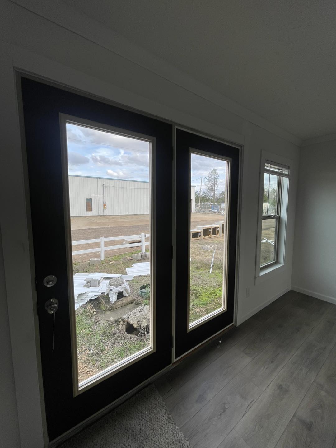 Modern room with wooden flooring and large glass doors opening to an outdoor construction scene. Overcast day, minimalist interior, calm ambiance.