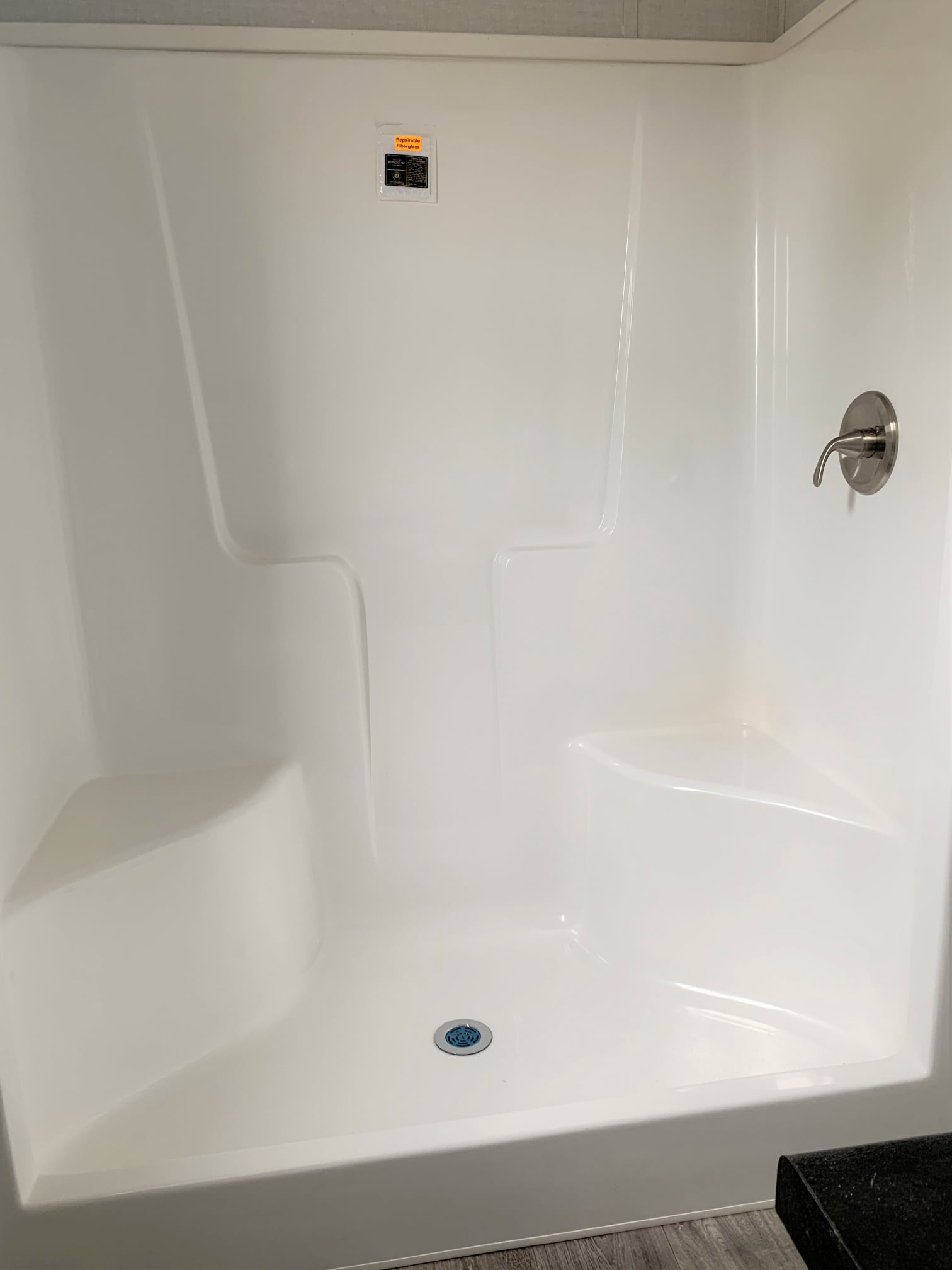 White shower stall with built-in corner seats and a single silver faucet on the right side. The floor is light wood, giving a clean, modern feel.