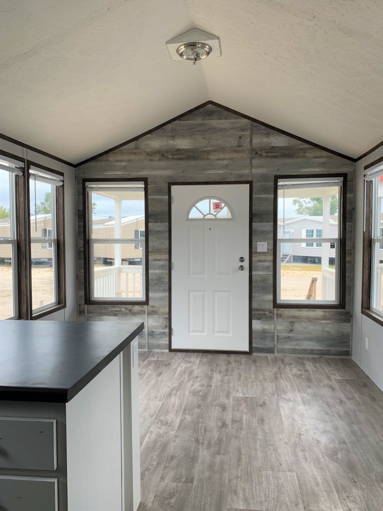 Bright room with a sloped ceiling, featuring a white door flanked by large windows. The interior has gray wood accents and laminate flooring.