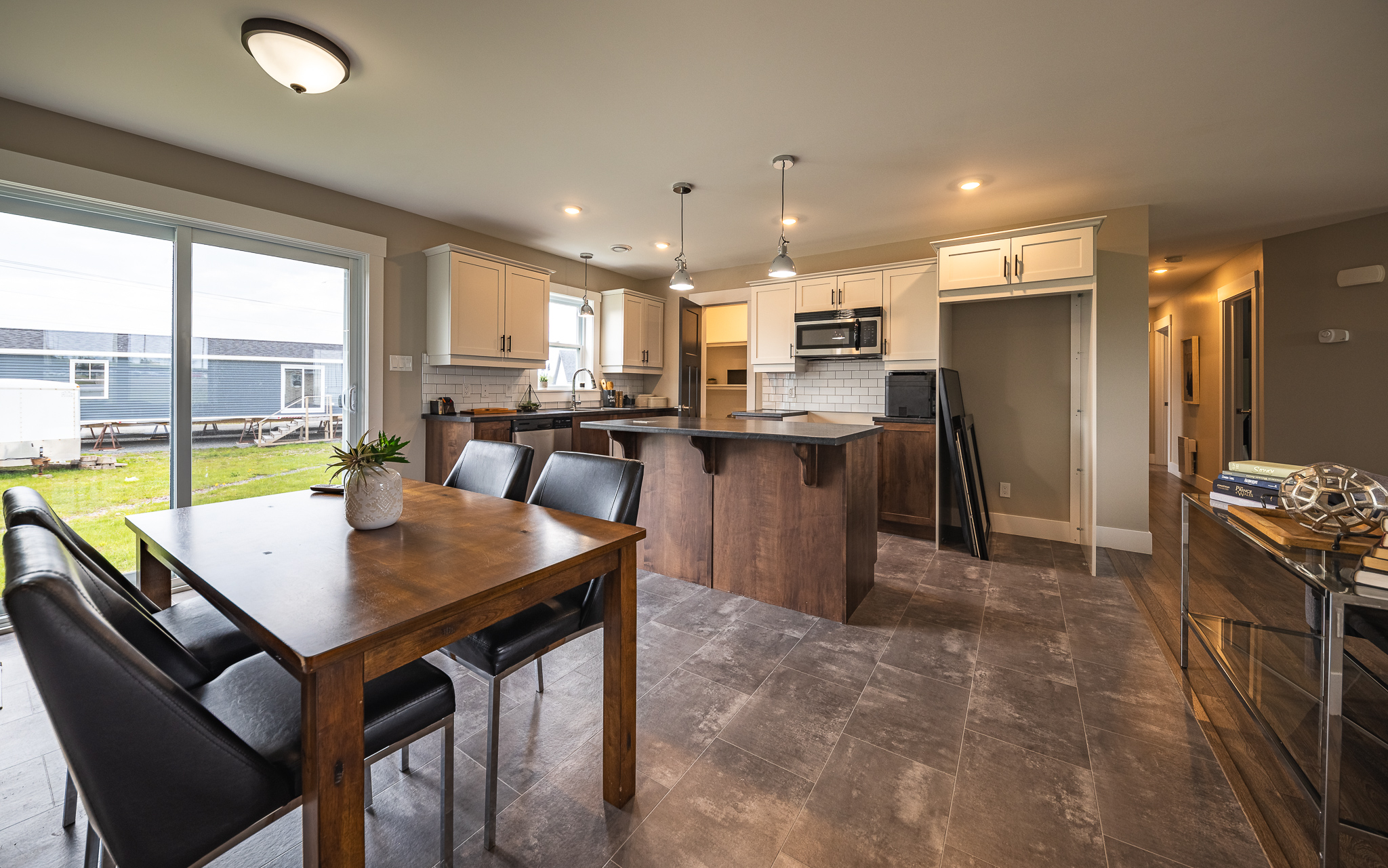 Modern kitchen and dining area with gray tiled floor, wooden cabinets, and a central island. Large windows provide natural light, creating a cozy, inviting atmosphere.