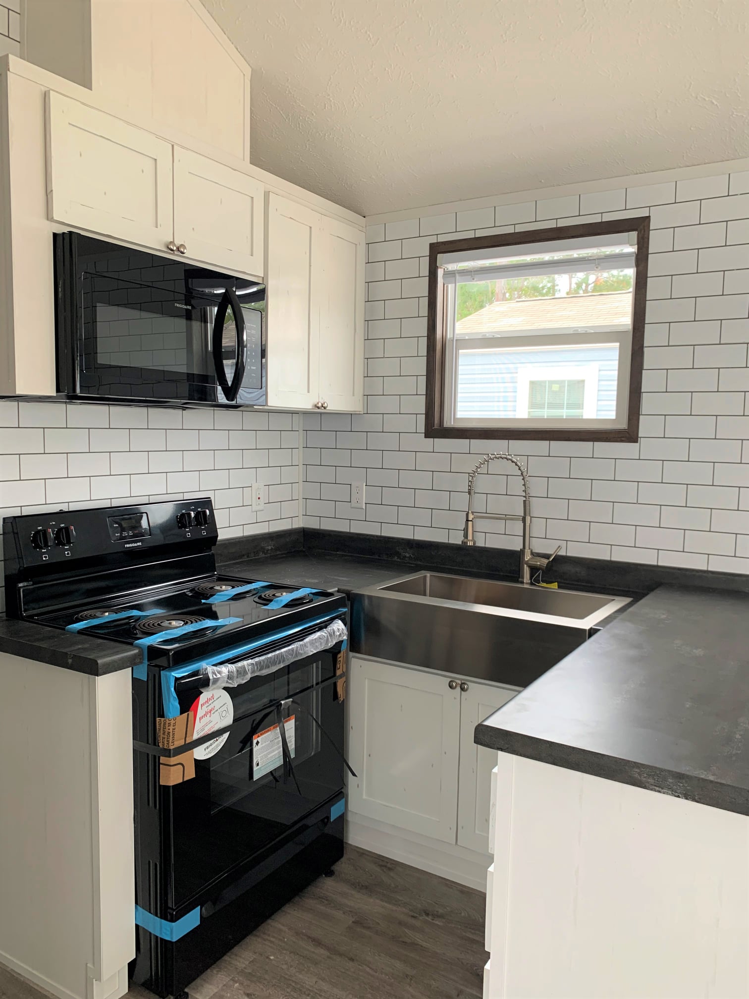 Compact kitchen with white cabinets, black countertops, and a window. Features a wrapped black oven, stainless steel sink, and white subway tiles for a modern look.