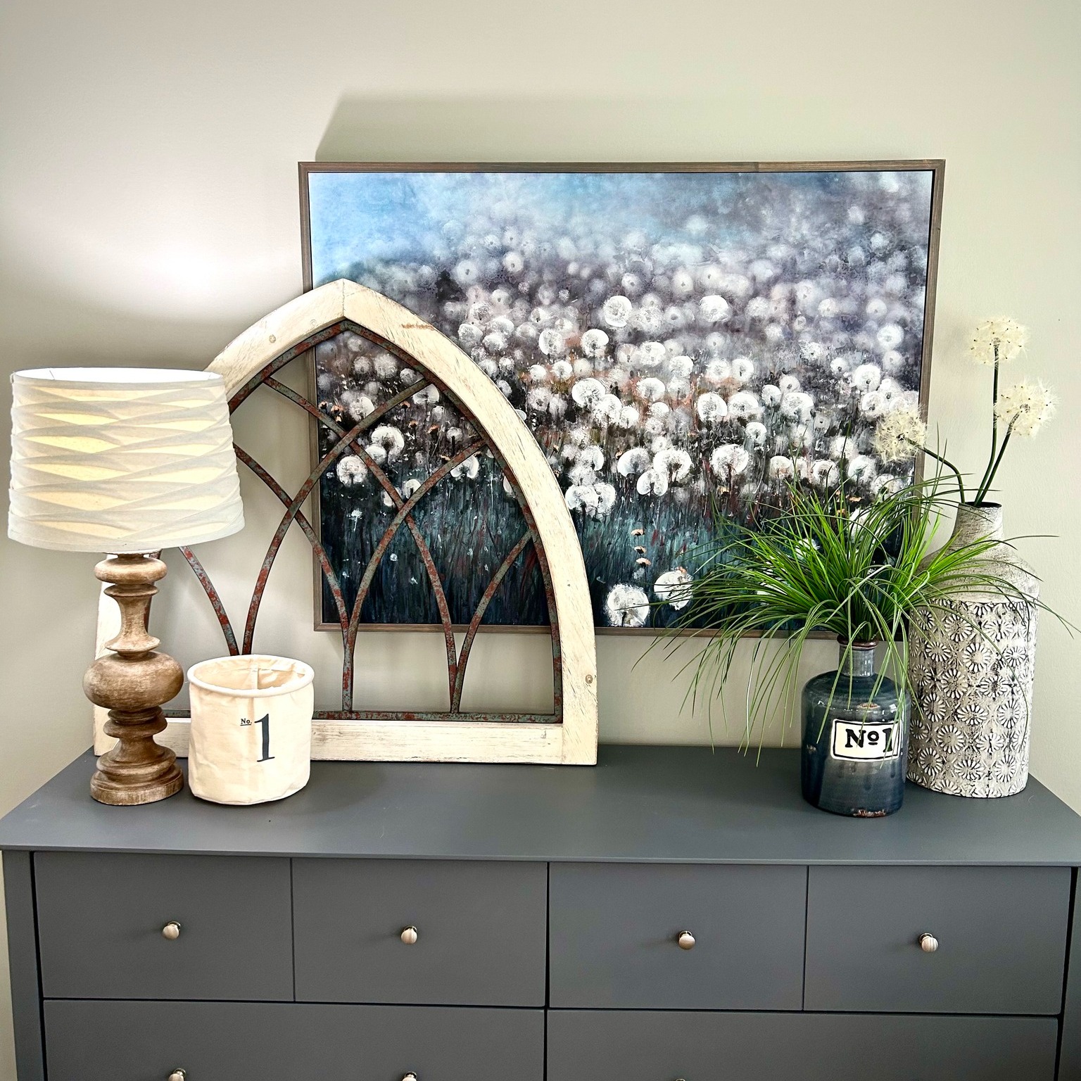 A stylish dresser topped with a lamp, vintage frame, numbered jars, and lush green plants. A floral painting in the background adds elegance.