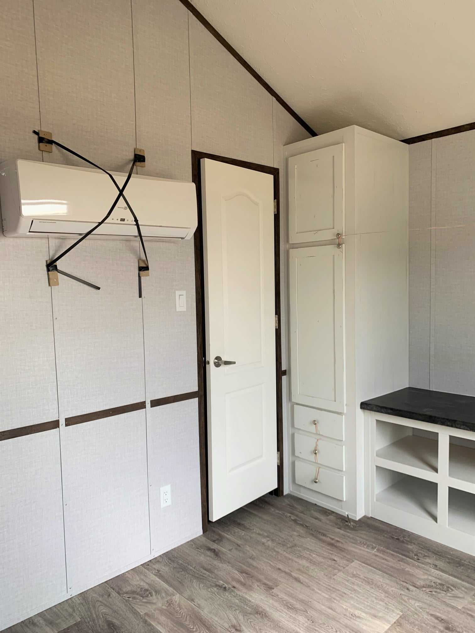 Minimalist room with light wood flooring; walls are gray with dark wood trim. A white door is flanked by an air conditioner and built-in shelves.