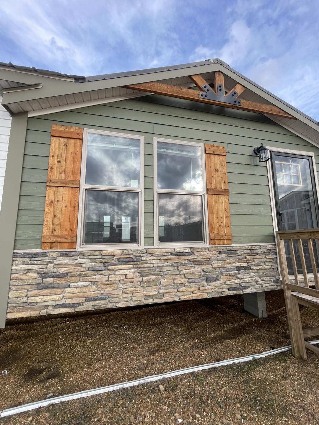 Green house facade with wooden shutters and stone base. The sky is cloudy, reflecting in the windows, creating a serene, rustic look.