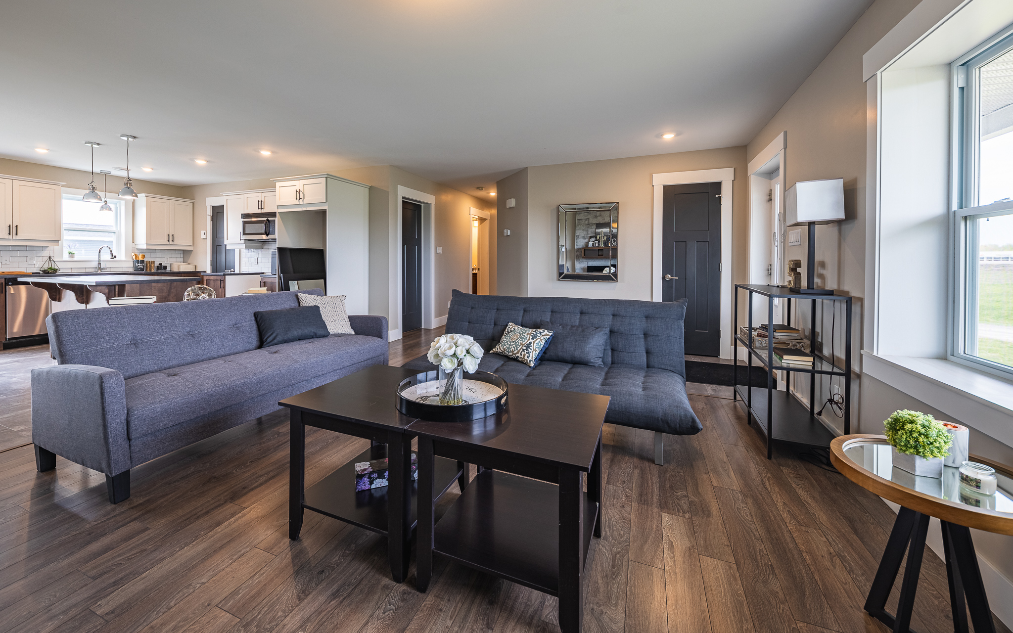 Modern living room with dark wood floors, gray sofas, and a black coffee table with flowers. Open kitchen in the background, bright and spacious.