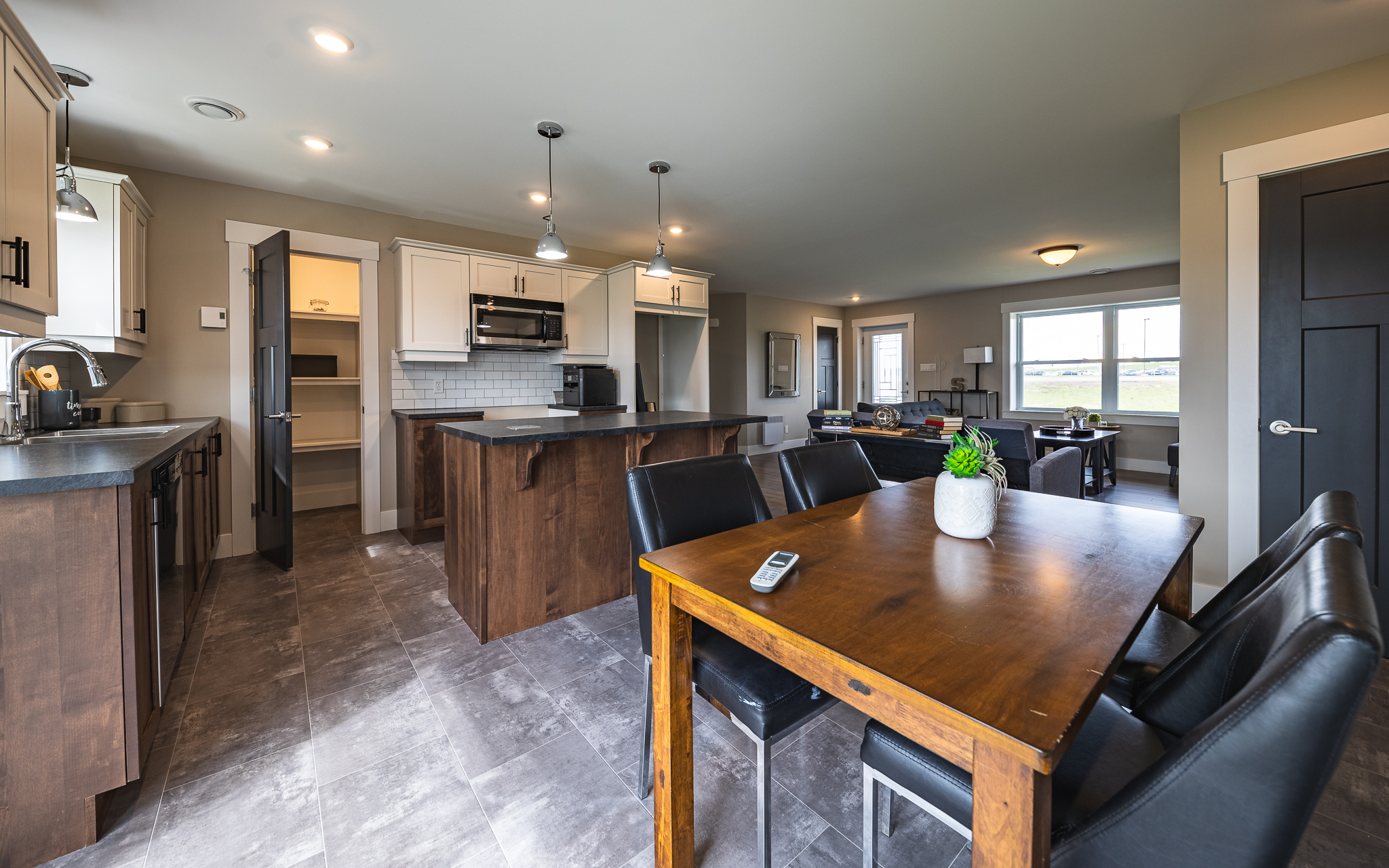 Spacious modern kitchen and dining area with wooden cabinets, a dark dining table with black chairs, pendant lights, and gray tiled floor. Bright and inviting.