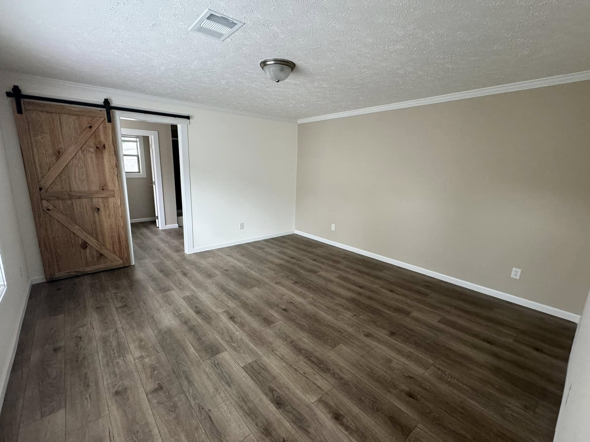 Spacious empty room with wood laminate flooring, a beige accent wall, and a wooden sliding barn door. The atmosphere is calm and minimalist.
