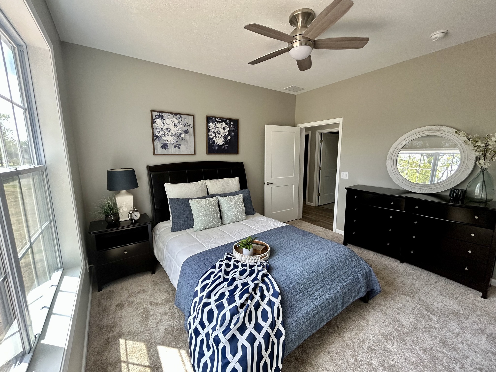 Bright bedroom with a large window, featuring a bed with navy and white bedding, dark furniture, and floral artwork. The room has a calm, cozy feel.