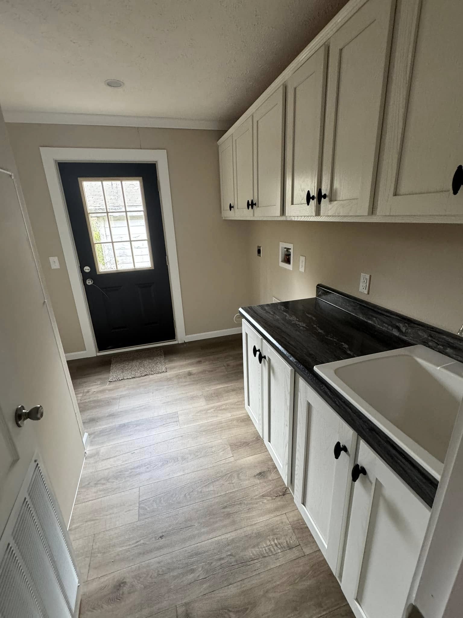 Narrow laundry room with wood floor, white cabinets, a dark countertop, and a deep sink. Black door at the end with a window, creating a minimalist feel.