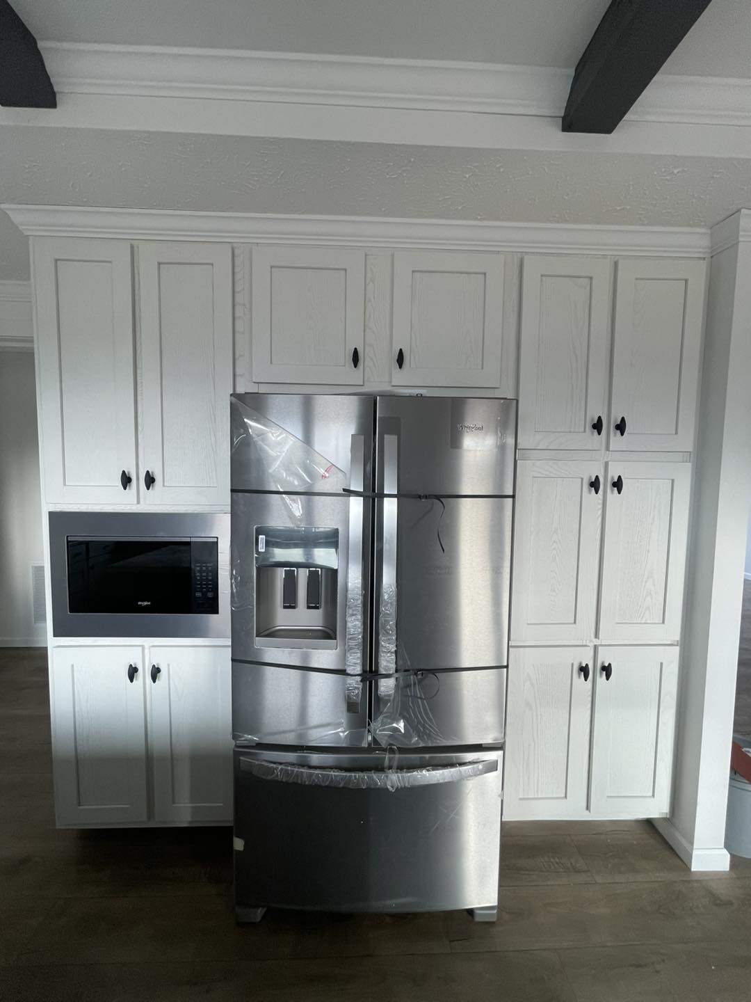 Stainless steel refrigerator with protective plastic stands against white cabinets, featuring a built-in water dispenser, beside a microwave. Modern kitchen scene.